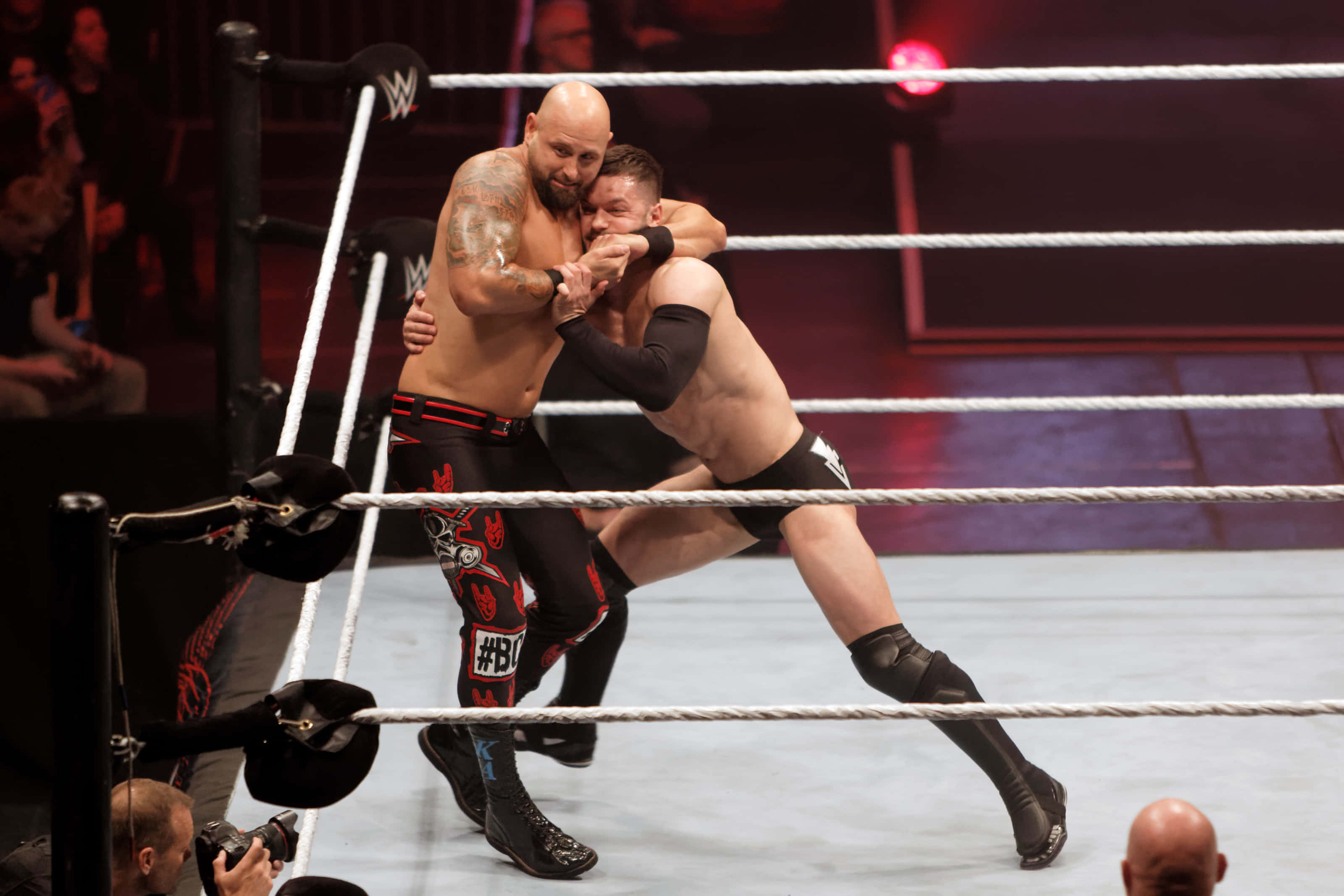WWE 桌布 壁紙 — 兩名肌肉發達的摔角手在燈光明亮的擂台上激烈糾纏，背景有繩索和人群。