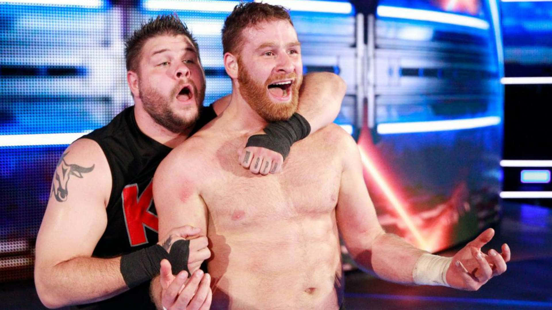 sami zayn 桌布 壁紙 — 兩名肌肉發達的摔角手在燈光璀璨的擂台上戲劇性地擁抱，藍色和紅色的燈光交織。
