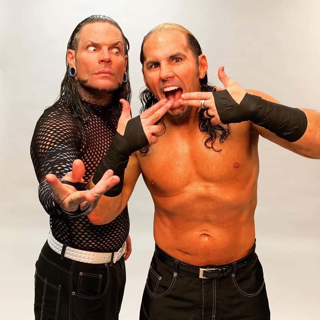 WWE 桌布 壁紙 — 兩名肌肉發達的摔角手在純白色背景前擺出戲劇性的姿勢，一人赤裸上身戴著黑色臂套，另一人穿著黑色網眼上衣。