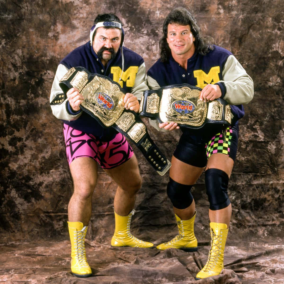 Rick Steiner 桌布 壁紙 — 兩名肌肉發達的摔角手穿著密西根主題外套和鮮豔短褲，在紋理棕色背景前自豪地展示冠軍腰帶。