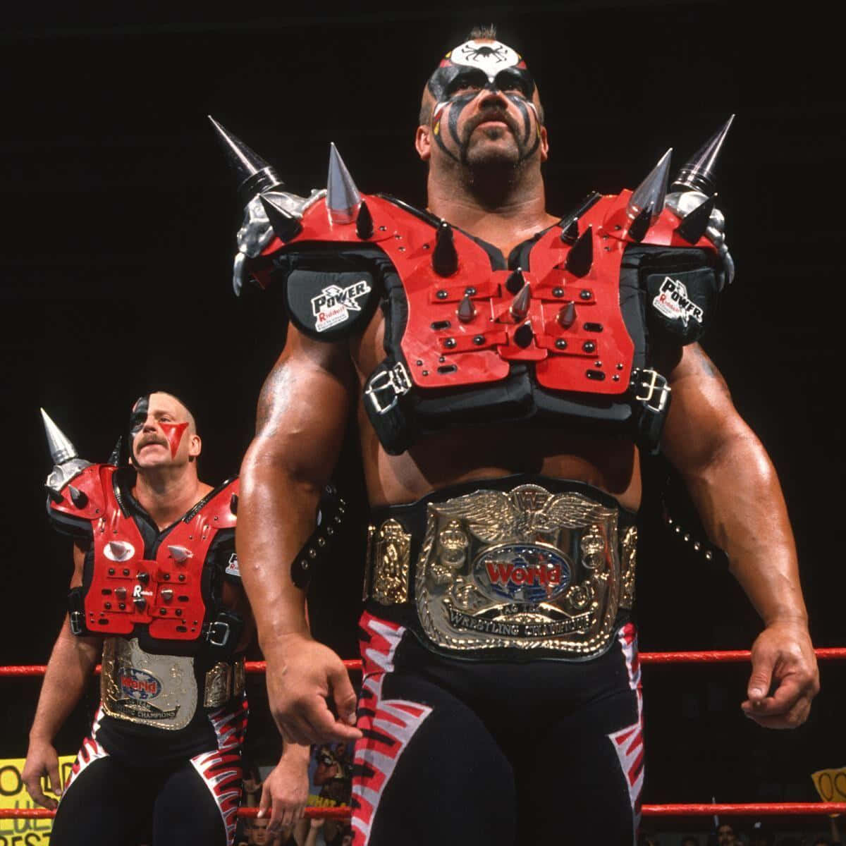 Road Warrior Hawk 桌布 壁紙 — 兩名肌肉發達的摔角手身穿紅色尖刺盔甲和冠軍腰帶，站在明亮的燈光下的摔角擂台上。