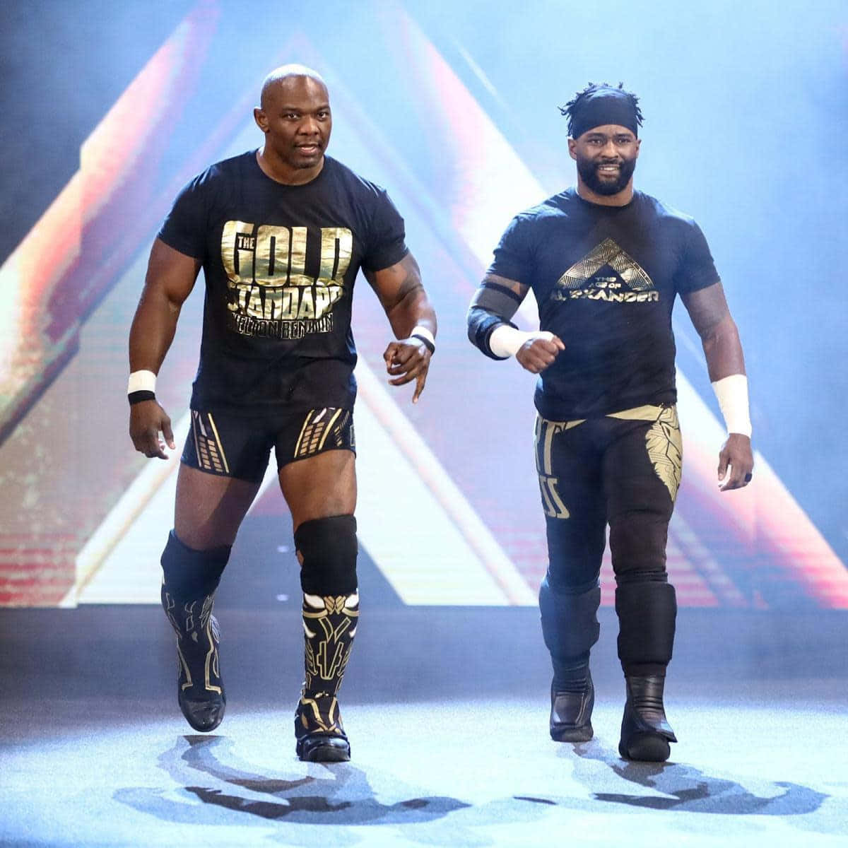 WWE 桌布 壁紙 — 兩名肌肉發達的摔角手身穿黑色和金色裝備，自信地走過一個明亮舞台，身後有幾何光束。