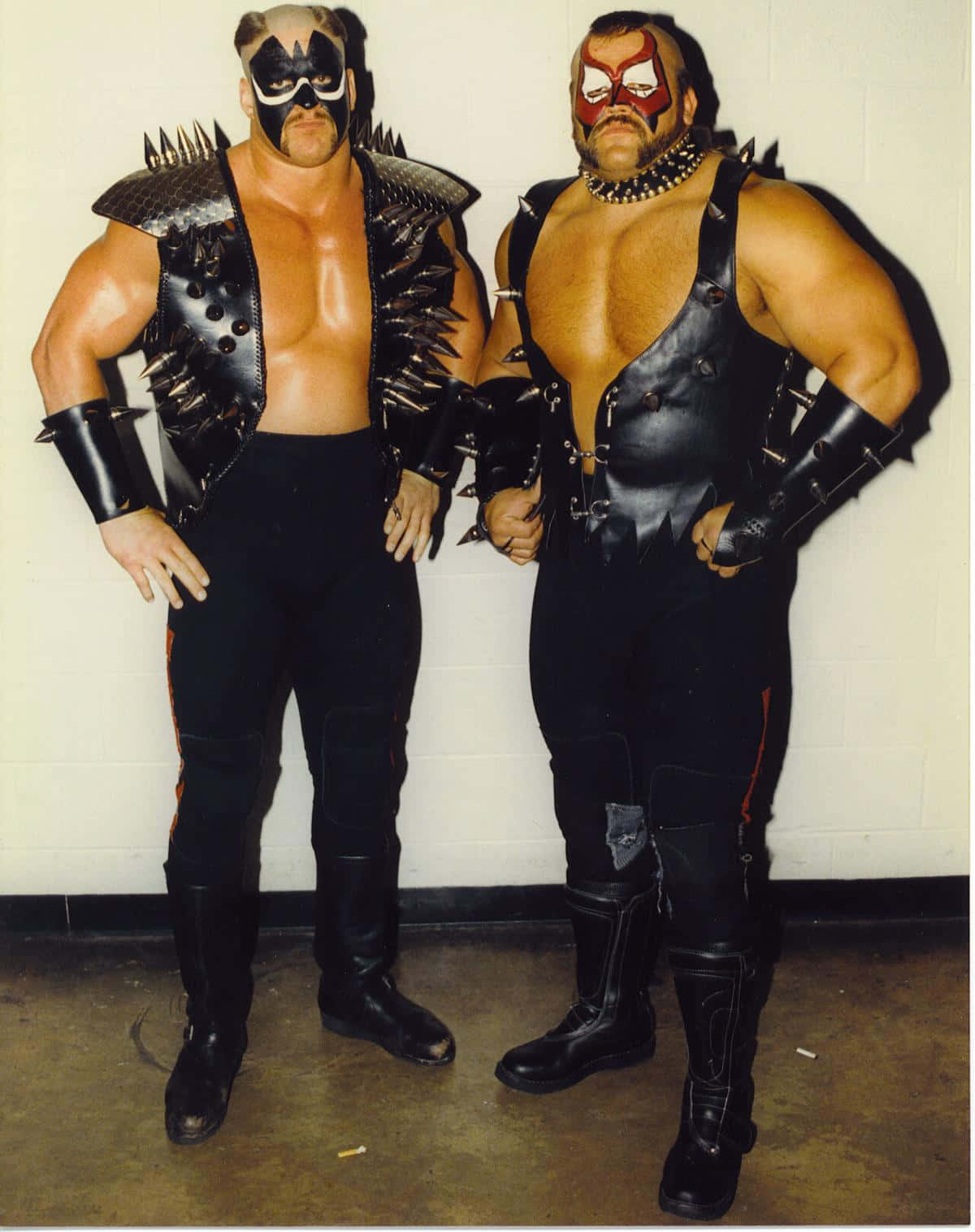 Road Warrior Hawk 桌布 壁紙 — 兩名肌肉發達的摔角手身穿黑色釘皮裝備和臉部彩繪，自信地靠著素牆擺姿勢。