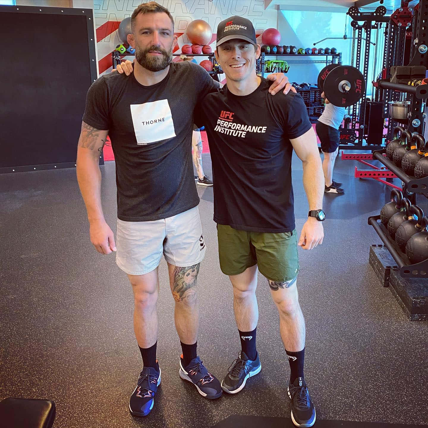 MMA 桌布 壁紙 — 兩名肌肉發達的男子在設備齊全、佈滿舉重和健身器材的健身房中互相搭肩擺姿勢。