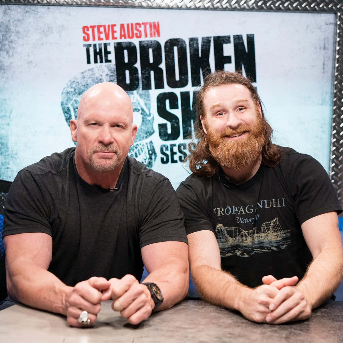 sami zayn 桌布 壁紙 — 兩名肌肉發達的男子坐在一張桌子前，背景寫著「Steve Austin The Broken Skull Sessions」，表情嚴肅且微笑。