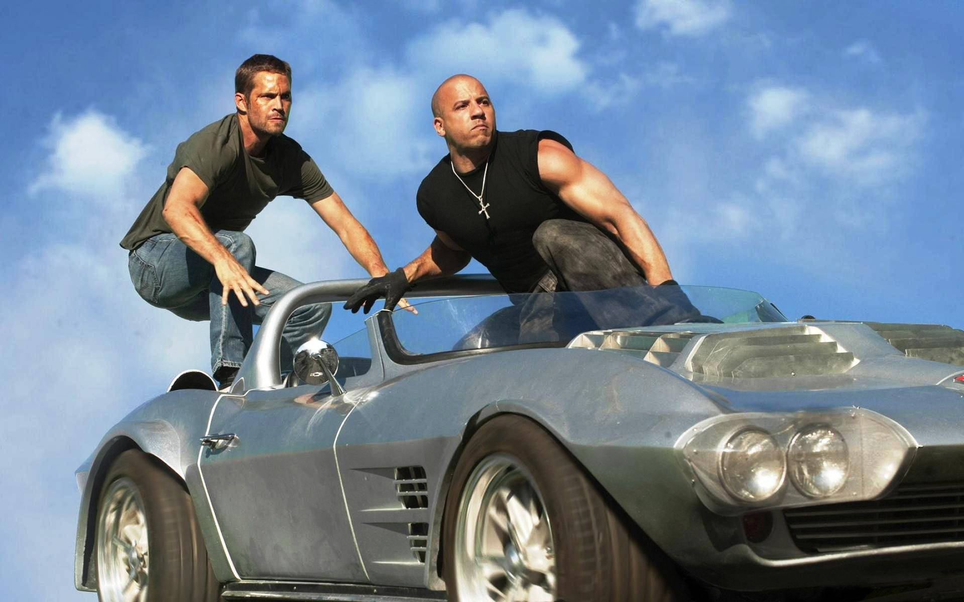 Paul Walker 桌布 壁紙 — 兩名肌肉發達的男子從一輛疾馳的銀色跑車中探出身子，背景是晴朗的藍天和白雲。