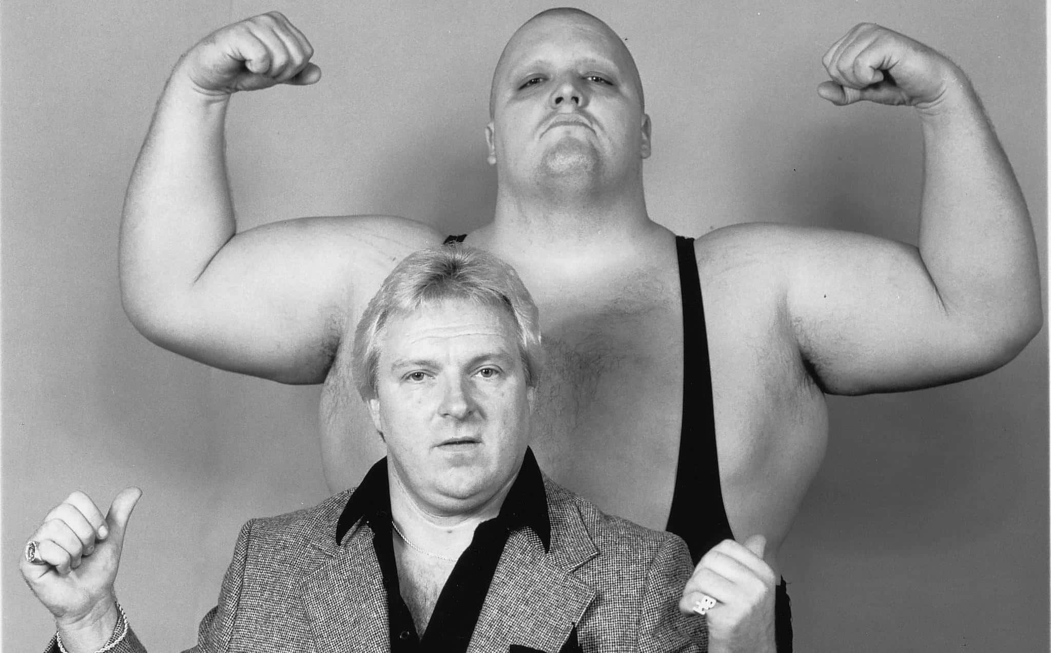 Bobby Heenan 桌布 壁紙 — 兩名肌肉發達的男子戲劇性地擺姿勢，一人身穿西裝坐著，另一人站在後面彎曲手臂，黑白攝影棚拍攝。