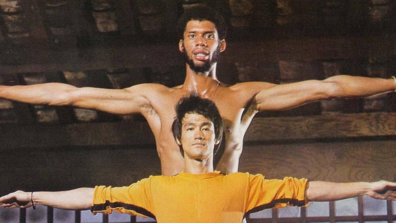 Kareem 桌布 壁紙 — 兩名肌肉發達的男子擺出同步的張臂姿勢，一人赤裸上身，另一人穿著黃色襯衫，背景為質樸的木質背景。