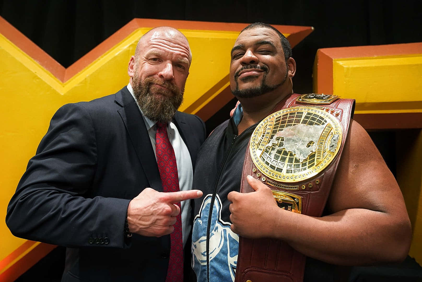 WWE 桌布 壁紙 — 兩名肌肉發達的男子自信地擺姿勢，一人身穿西裝指向另一人，後者自豪地舉著冠軍腰帶，背景為大膽的黃色和橙色。