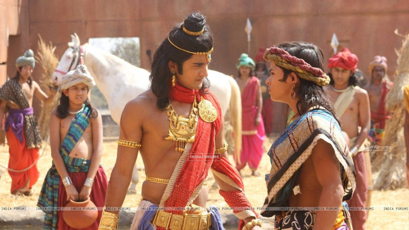 Indian 桌布 壁紙 — 兩名肌肉發達的男子身穿華麗的古代印度戰士服裝，在塵土飛揚的村莊廣場上對峙，背景有旁觀者和一匹白馬。