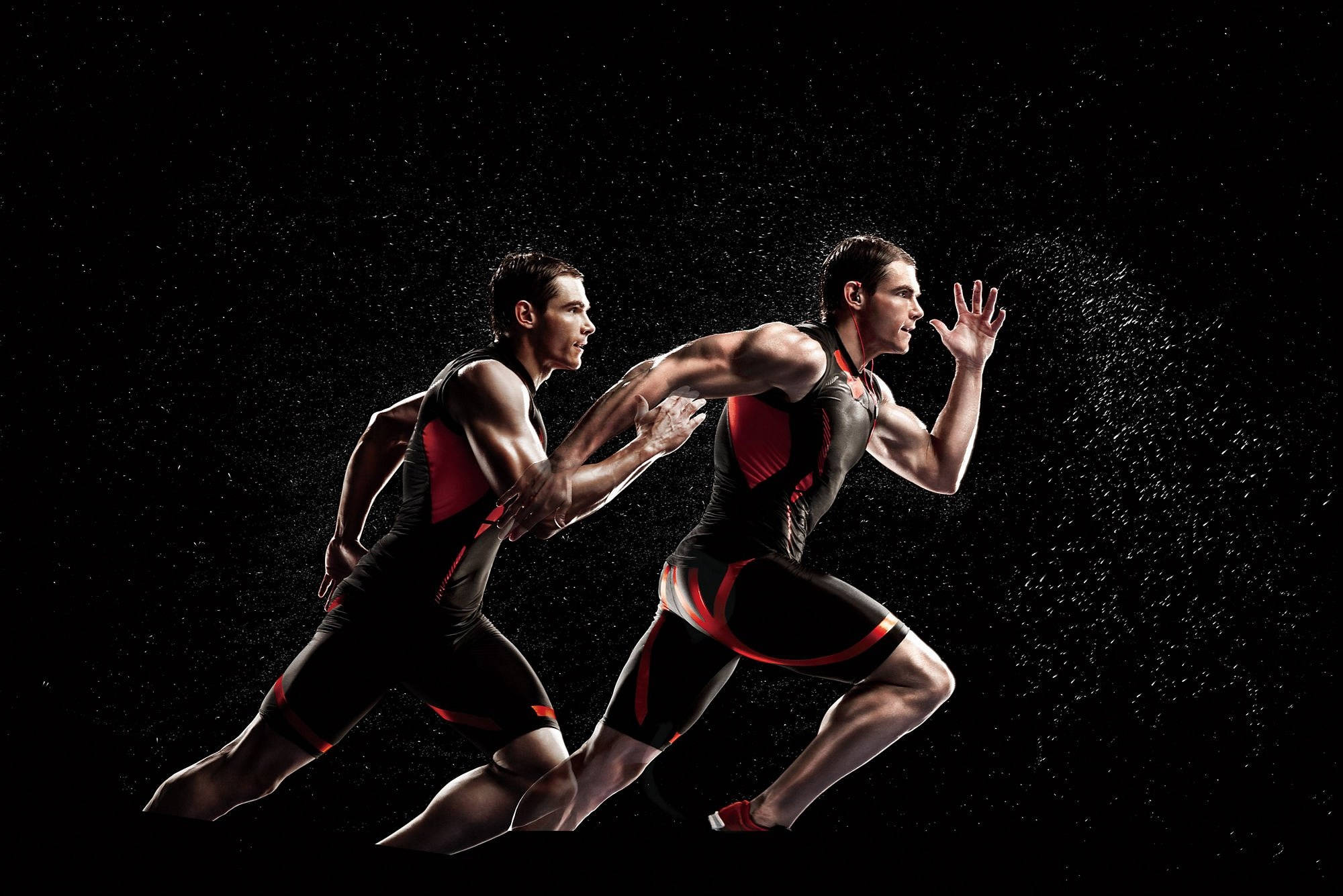 Athletic 桌布 壁紙 — 兩名肌肉發達的運動員穿著黑色和紅色的三項鐵人服，在純黑色背景前動態衝刺，水滴在他們周圍飛濺。