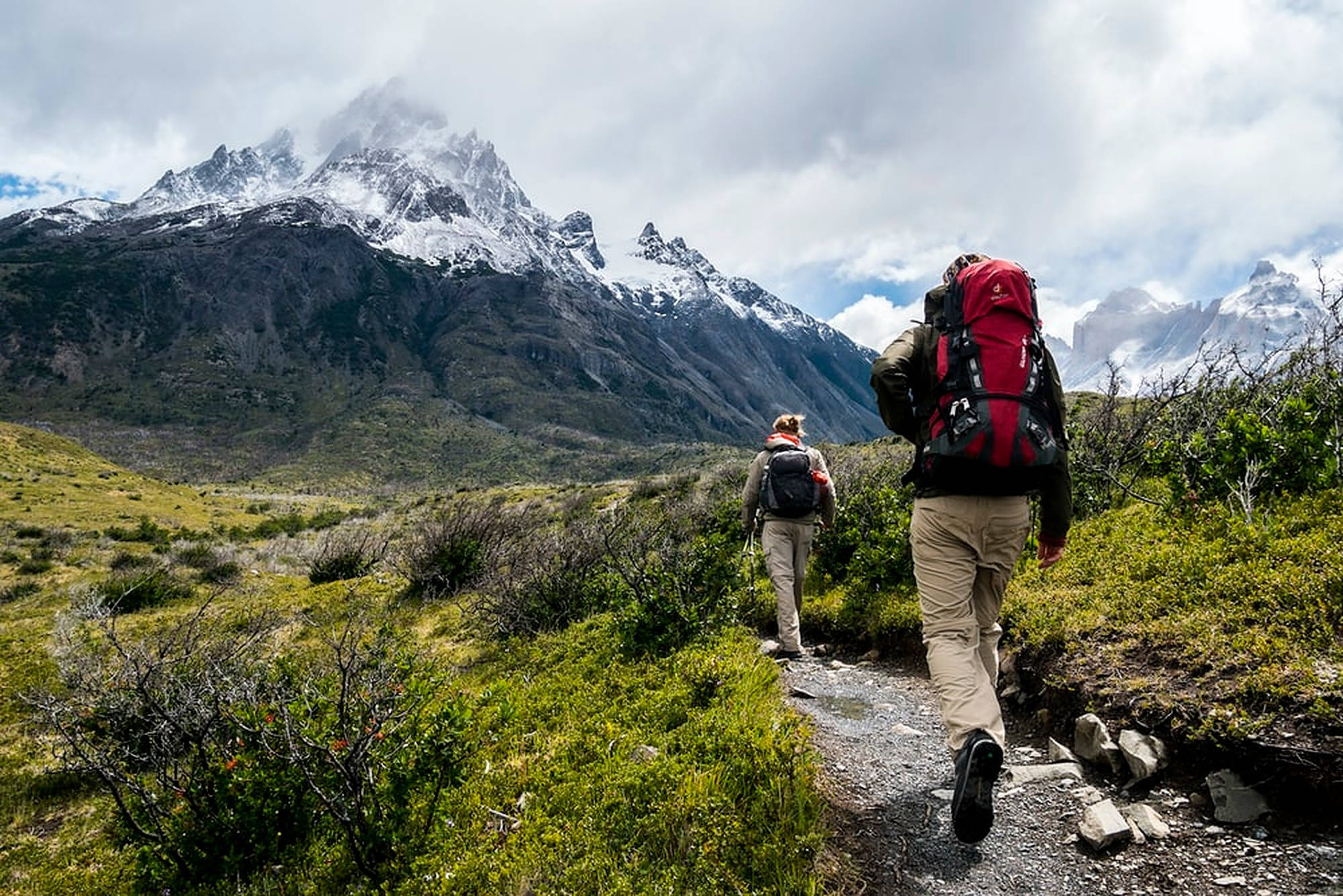 Hiking 桌布 壁紙 — 兩名背著大型背包的遠足者沿著一條岩石小徑穿過綠色灌木叢，在陰天之下向著積雪的山峰前進。