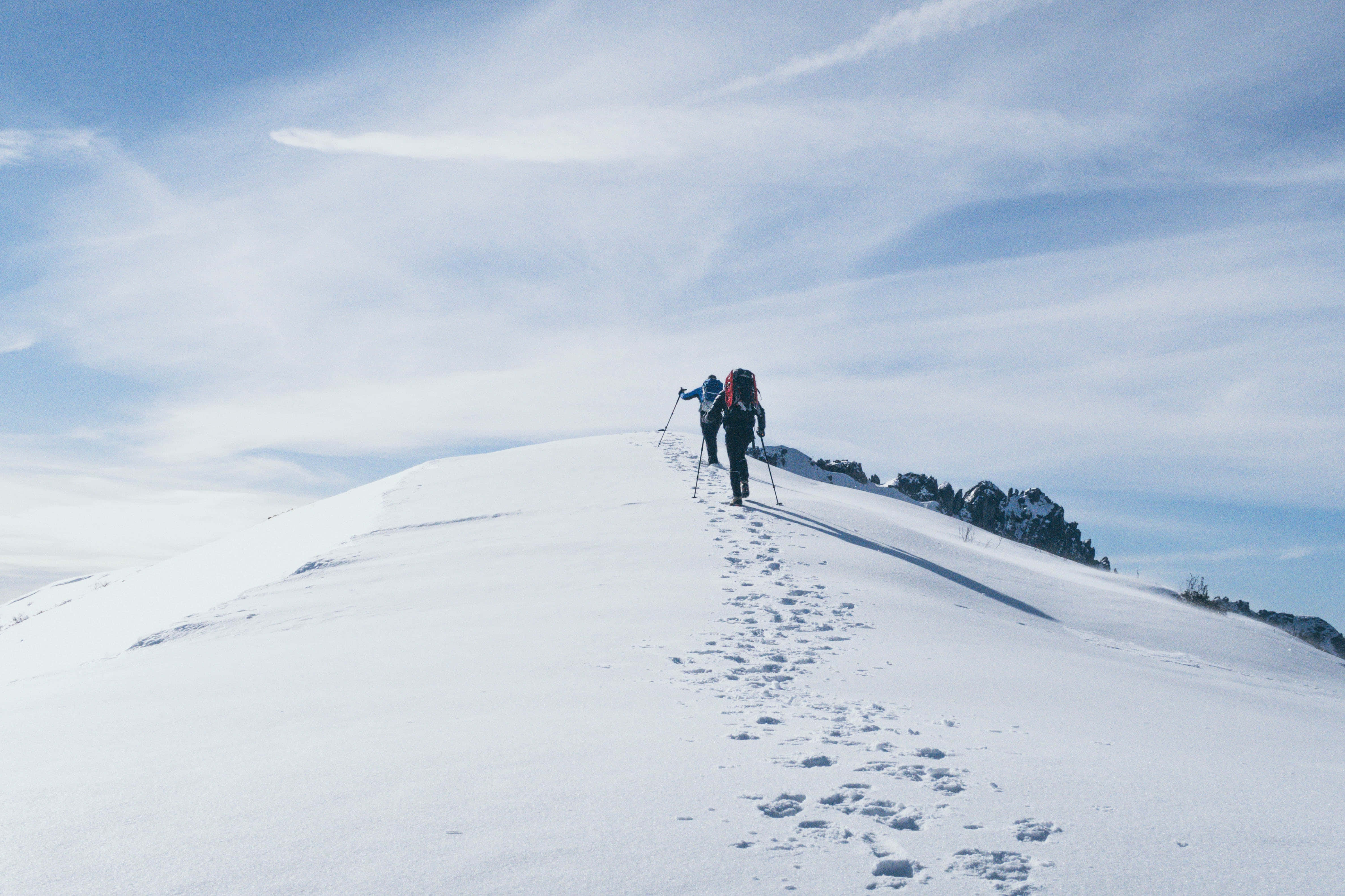 雪 桌布 壁紙 — 兩名背著背包、拿著登山杖的健行者，在晴朗藍天與薄雲下，沿著積雪的山脊向上行進。