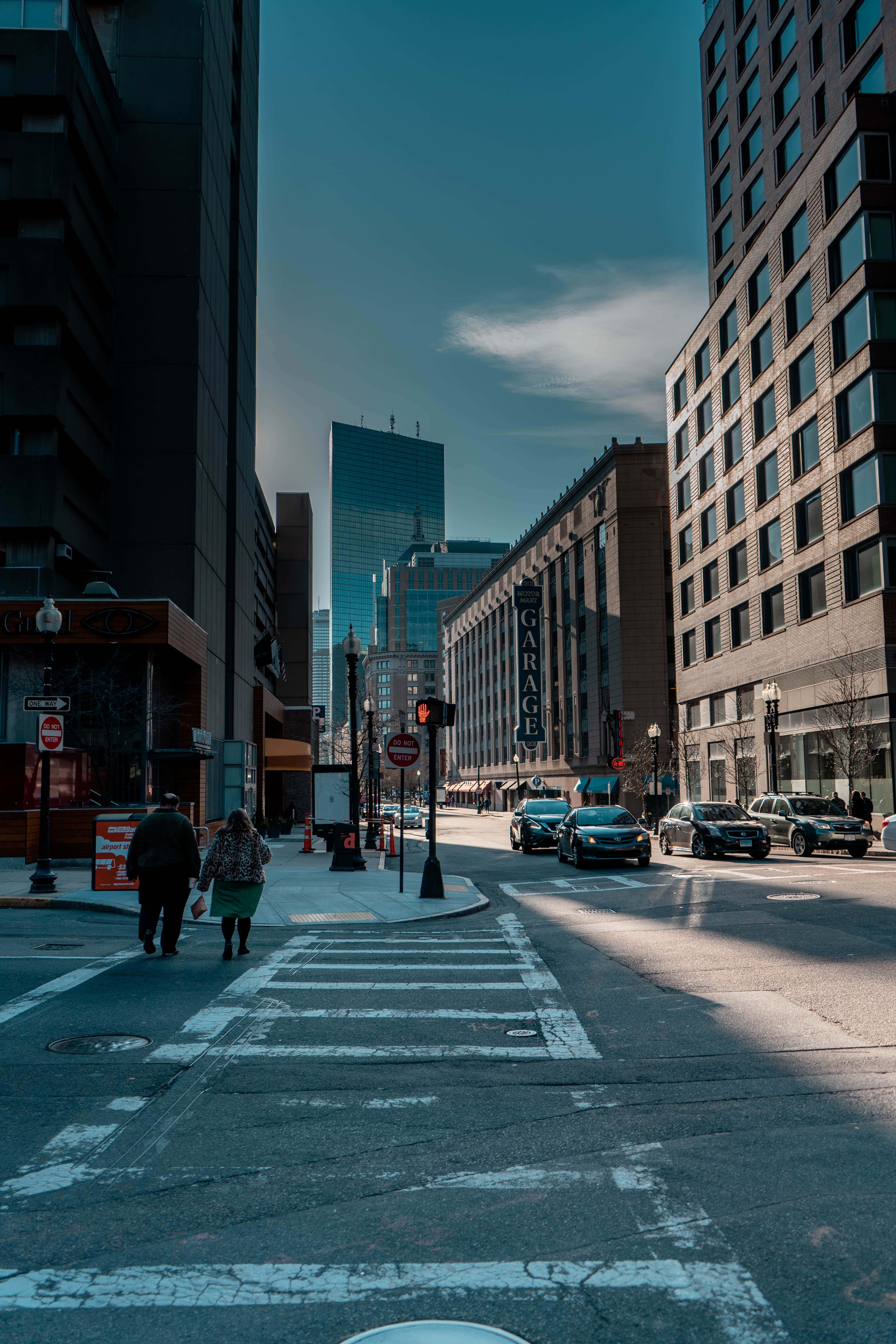 Sidewalk 桌布 壁紙 — 兩名行人正在陽光明媚的城市街道上過馬路，街道兩旁是高聳的摩天大樓和停泊的汽車，天空湛藍。