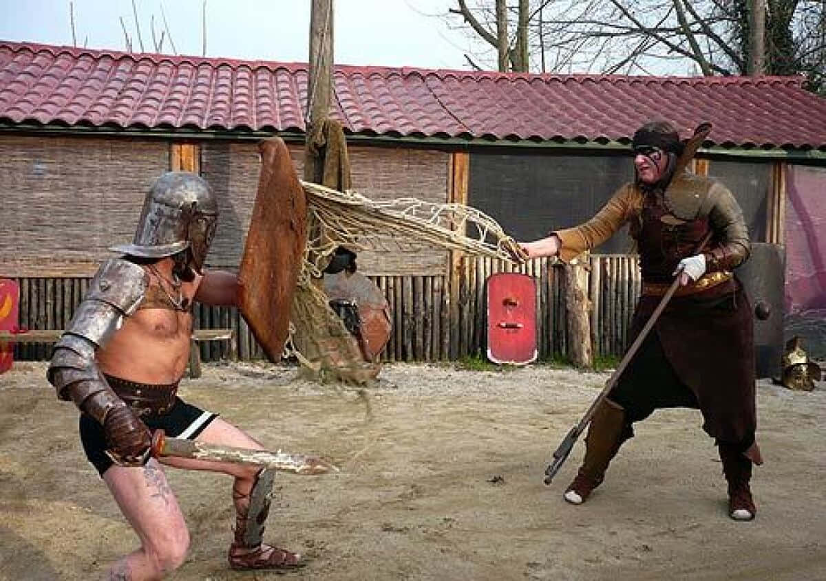 Gladiator 桌布 壁紙 — 兩名裝甲戰士在沙地競技場上交鋒，一人手持劍盾，另一人手持長矛和網，上方是紅瓦屋頂。