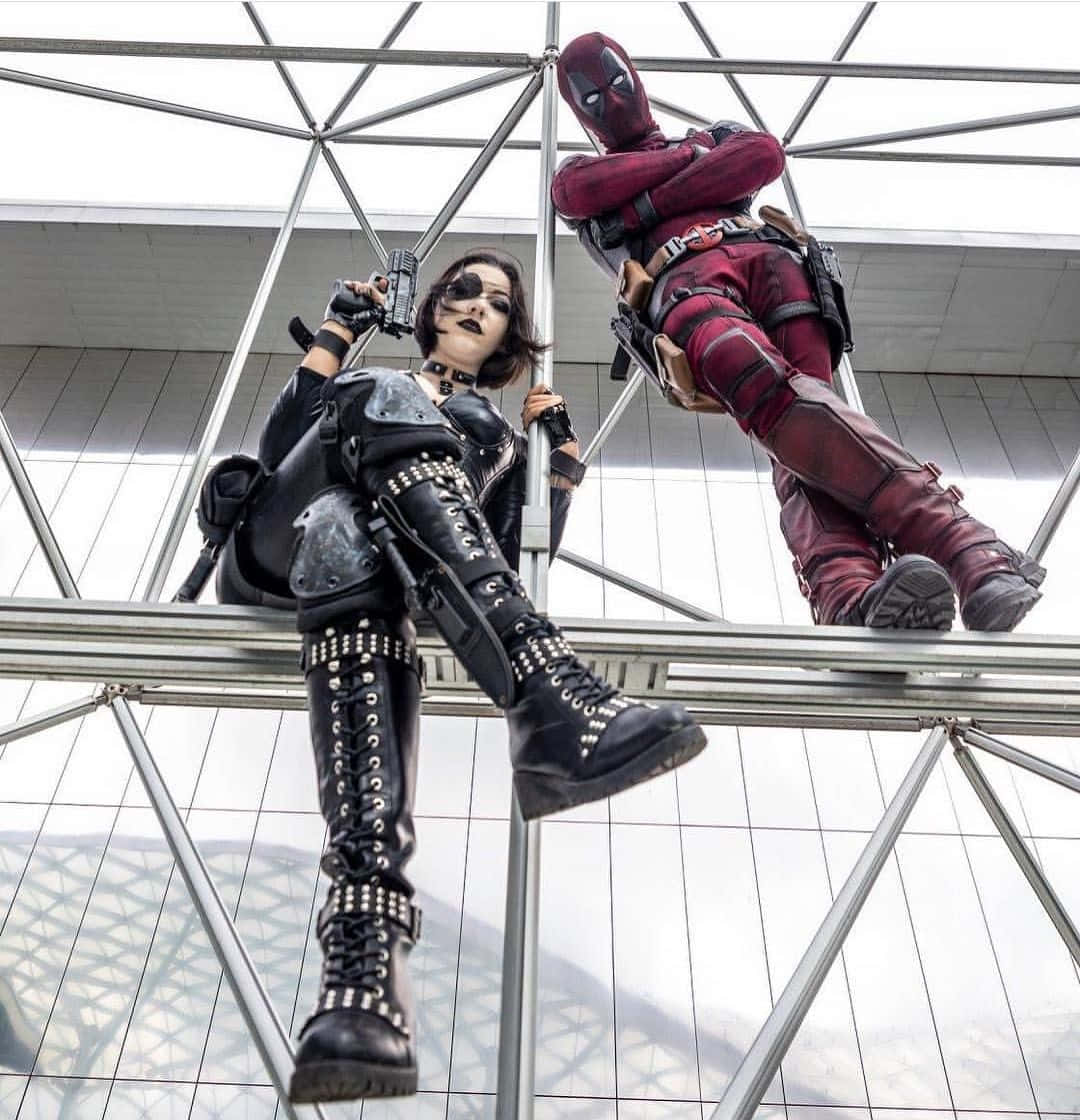 Deadpool And Domino 桌布 壁紙 — 兩名角色扮演者穿著死侍和黑寡婦服裝，在現代玻璃和鋼鐵屋頂結構上自信地擺姿勢。