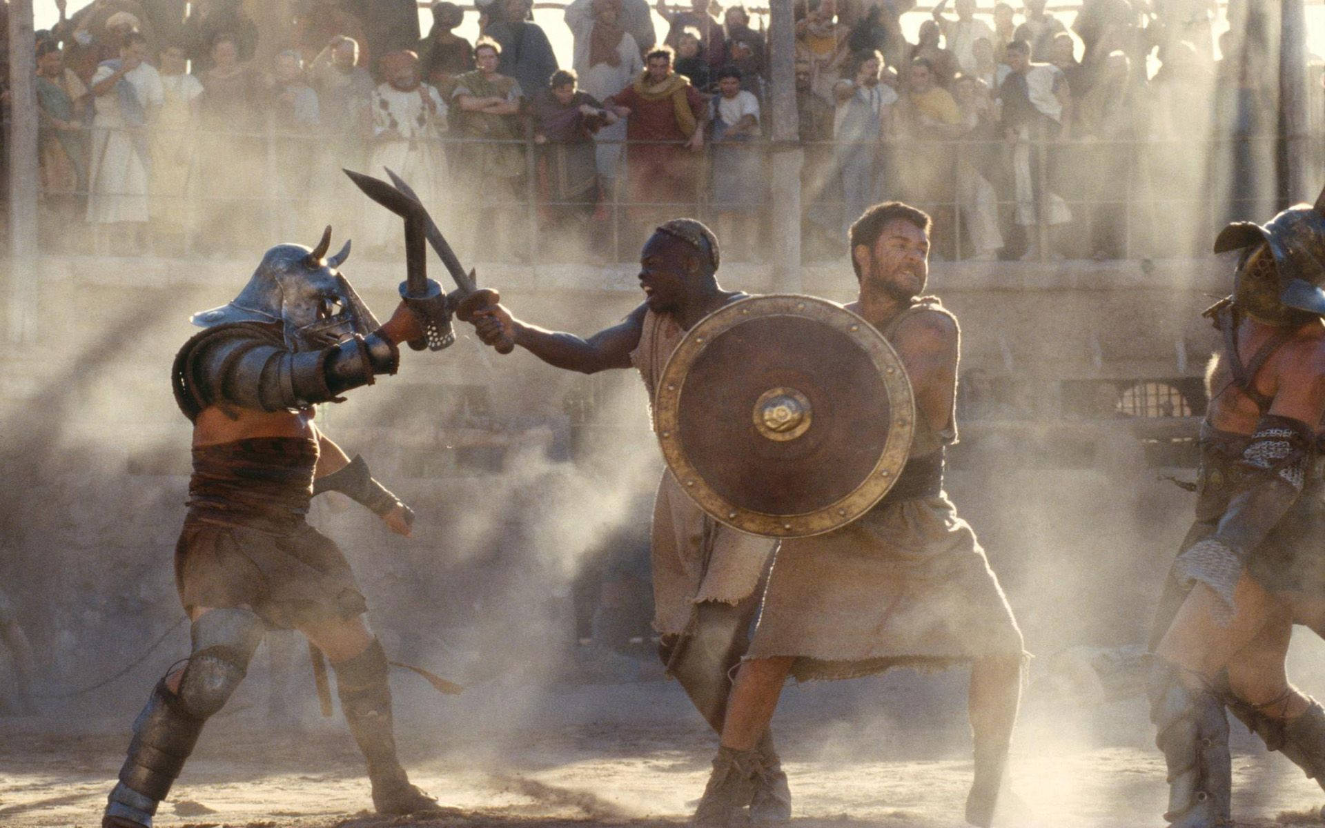 Gladiator 桌布 壁紙 — 兩名角鬥士在塵土飛揚的競技場中揮劍交鋒，觀眾在金色陽光下從石砌陽台觀看。