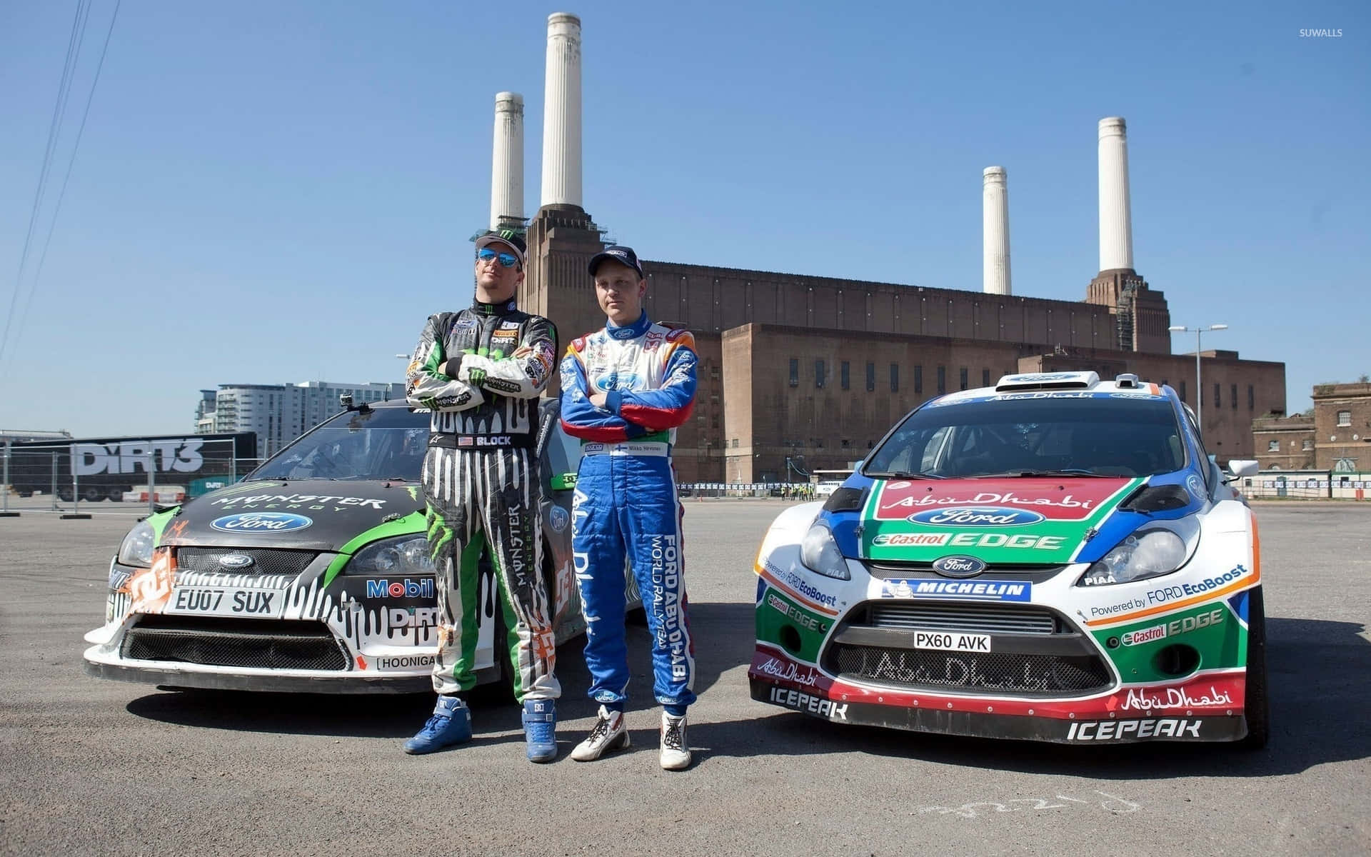 Ken Block 桌布 壁紙 — 兩名賽車手全副裝備，自信地站在他們的福特拉力賽車旁，背景是工業磚建築和高聳的煙囪，天空清澈湛藍。