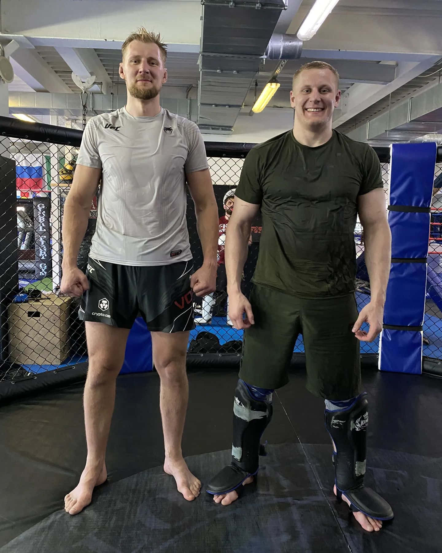 UFC 桌布 壁紙 — 兩名赤腳男性拳手在健身籠內，一人戴護脛，另一人穿運動裝備，兩人都對著鏡頭微笑。