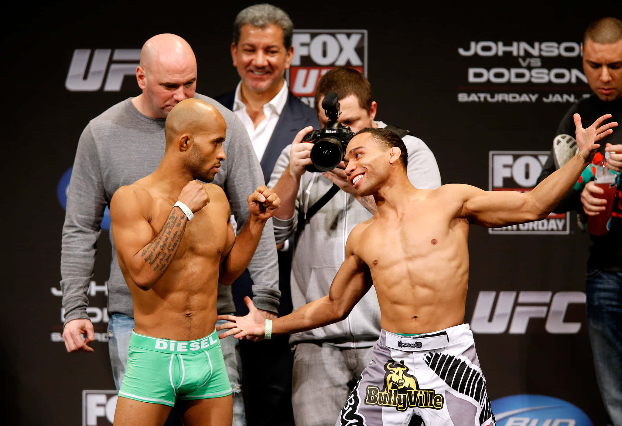 UFC 桌布 壁紙 — 兩名赤裸上身的UFC拳手在稱重儀式上對峙，身後閃光燈不斷，背景帶有品牌標誌。
