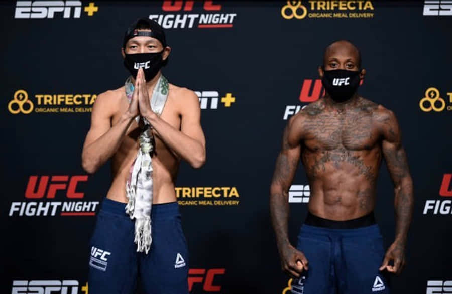 MMA 桌布 壁紙 — 兩名赤裸上身的UFC拳手站在過磅儀式上，一人雙手合十祈禱，另一人雙臂下垂，兩人都戴著黑色口罩和藍色短褲。