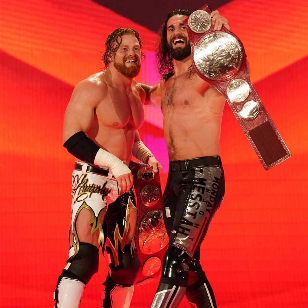 WWE 桌布 壁紙 — 兩名赤裸上身的摔角手在紅色燈光舞台上高舉冠軍腰帶慶祝勝利。