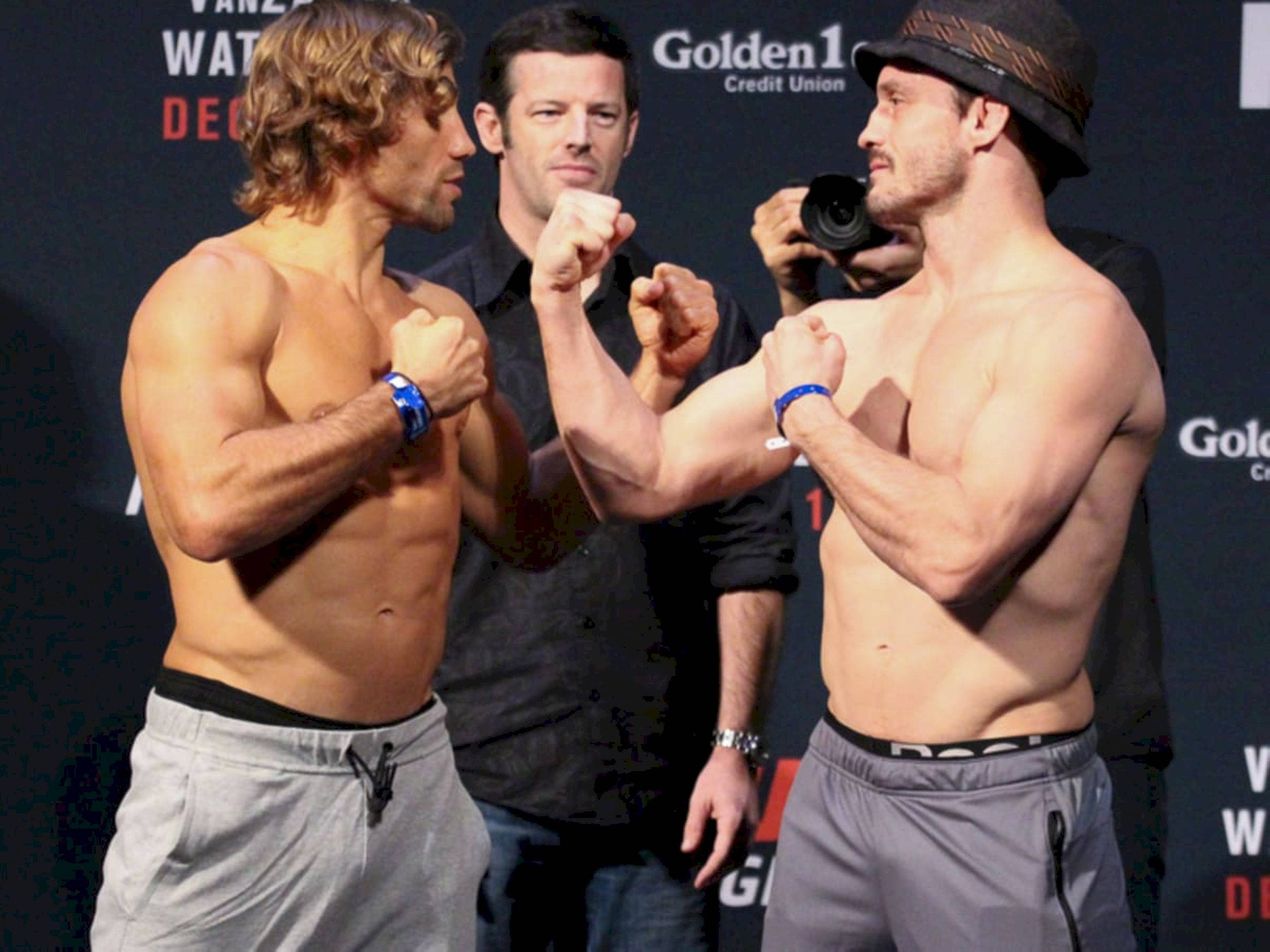 UFC 桌布 壁紙 — 兩名赤裸上身的男性拳手在稱重活動中進行緊張的賽前對峙，背景中有一名攝影師。