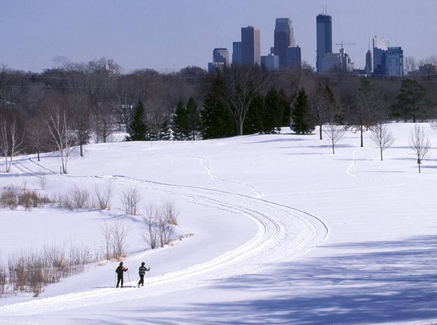 Minneapolis 桌布 壁紙 — 兩名越野滑雪者沿著蜿蜒的雪道滑行，遠處的城市天際線在光禿禿的樹木後方升起，天空湛藍清澈。