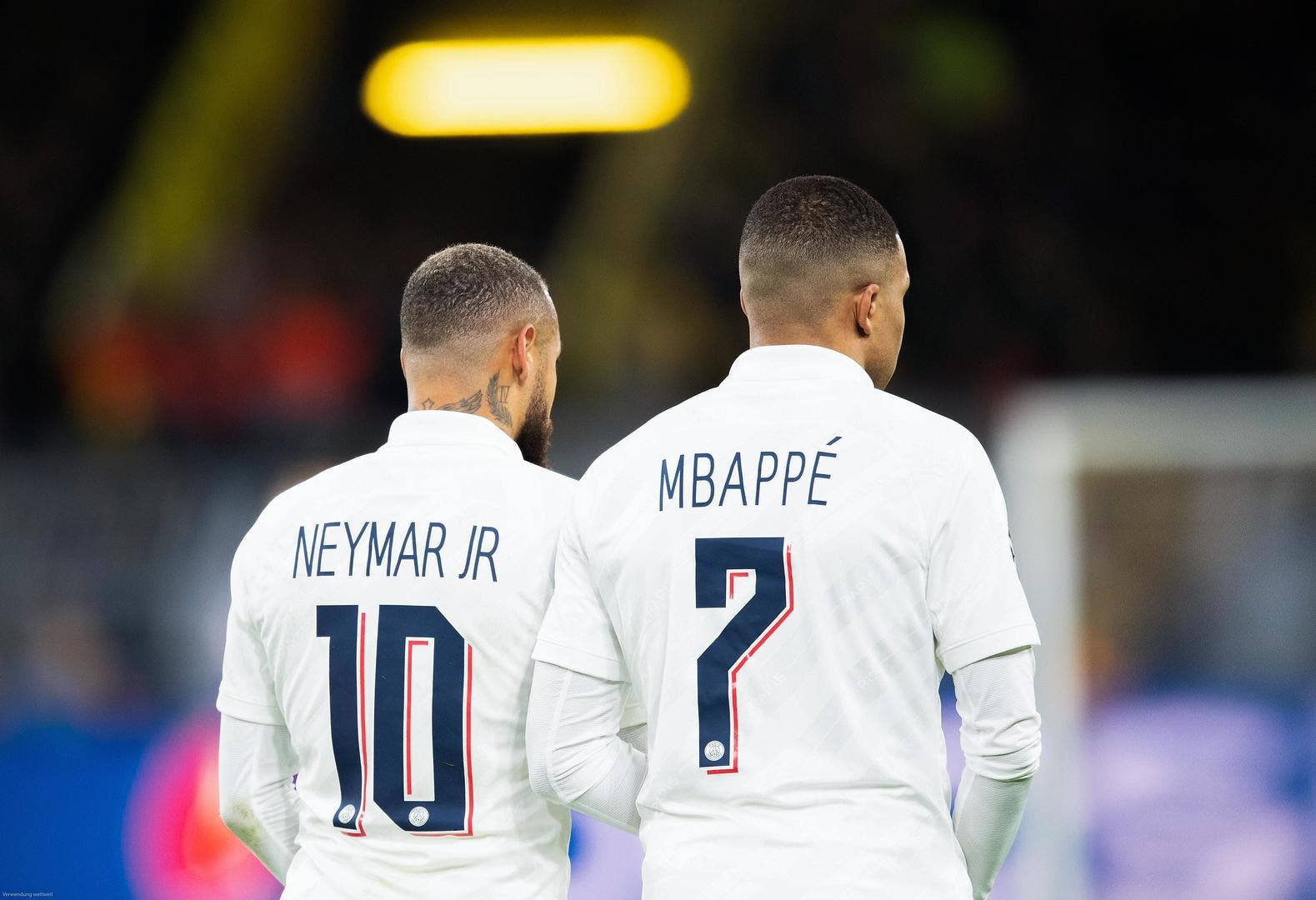 Kylian Mbappe 桌布 壁紙 — 兩名足球員內馬爾 Jr 和姆巴佩在燈光明亮的球場上背對背站立，身穿白色球衣，名字和號碼清晰可見。