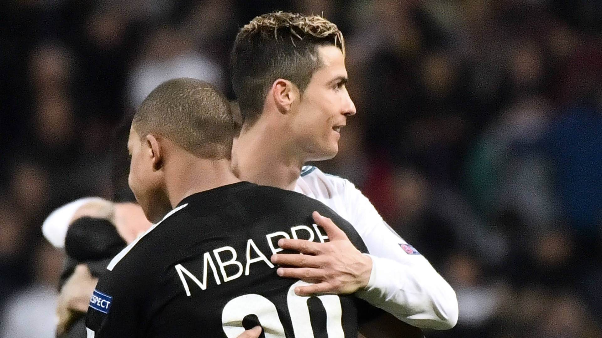 Kylian Mbappe 桌布 壁紙 — 兩名足球運動員在燈光明亮的球場上擁抱，一人身穿黑色球衣，上面有Mbarre 20字樣，另一人身穿白色球衣微笑