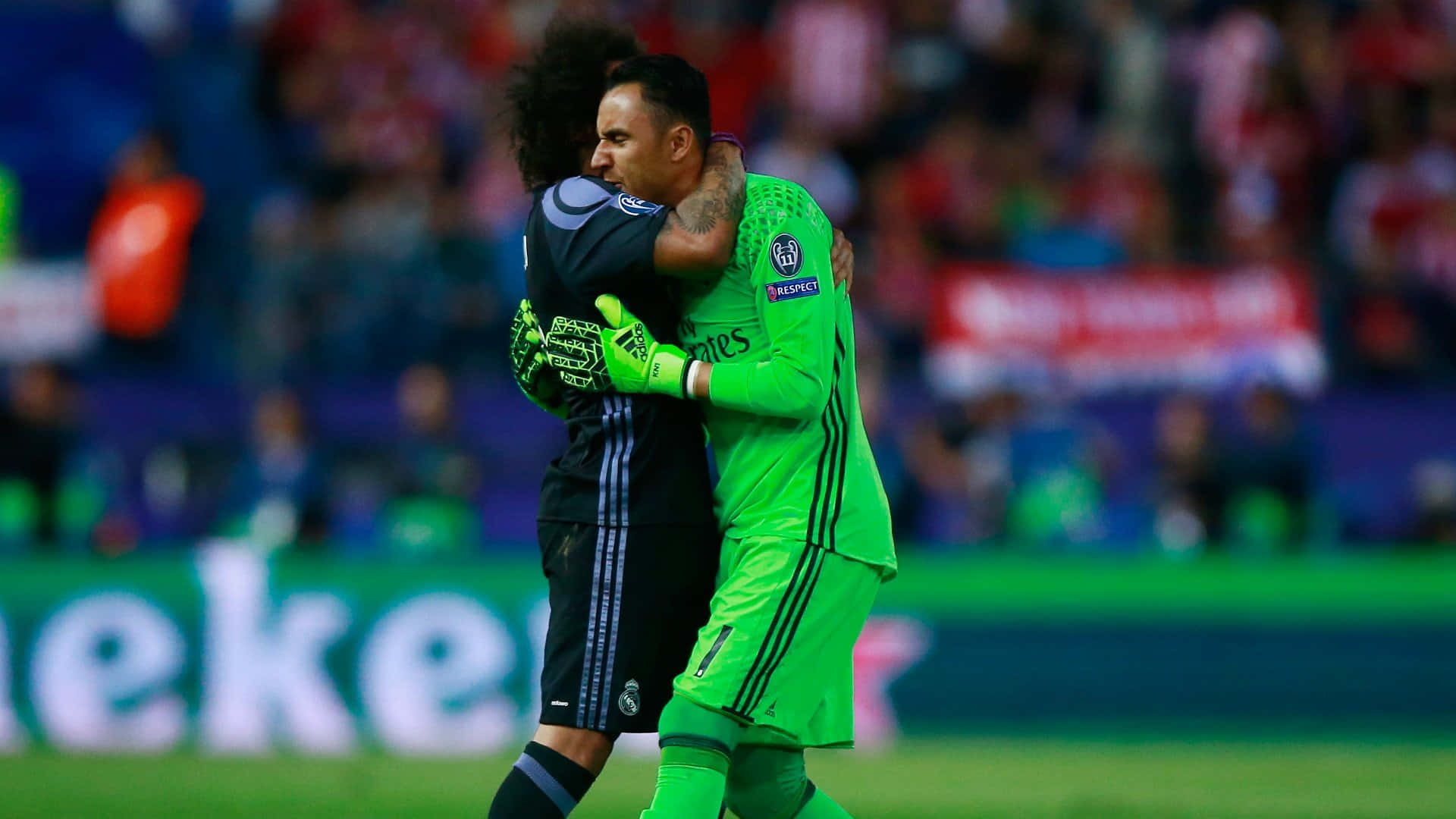 Keylor Navas 桌布 壁紙 — 兩名足球運動員在體育場燈光下，於充滿活力的綠色球場上擁抱，背景是模糊的球迷。