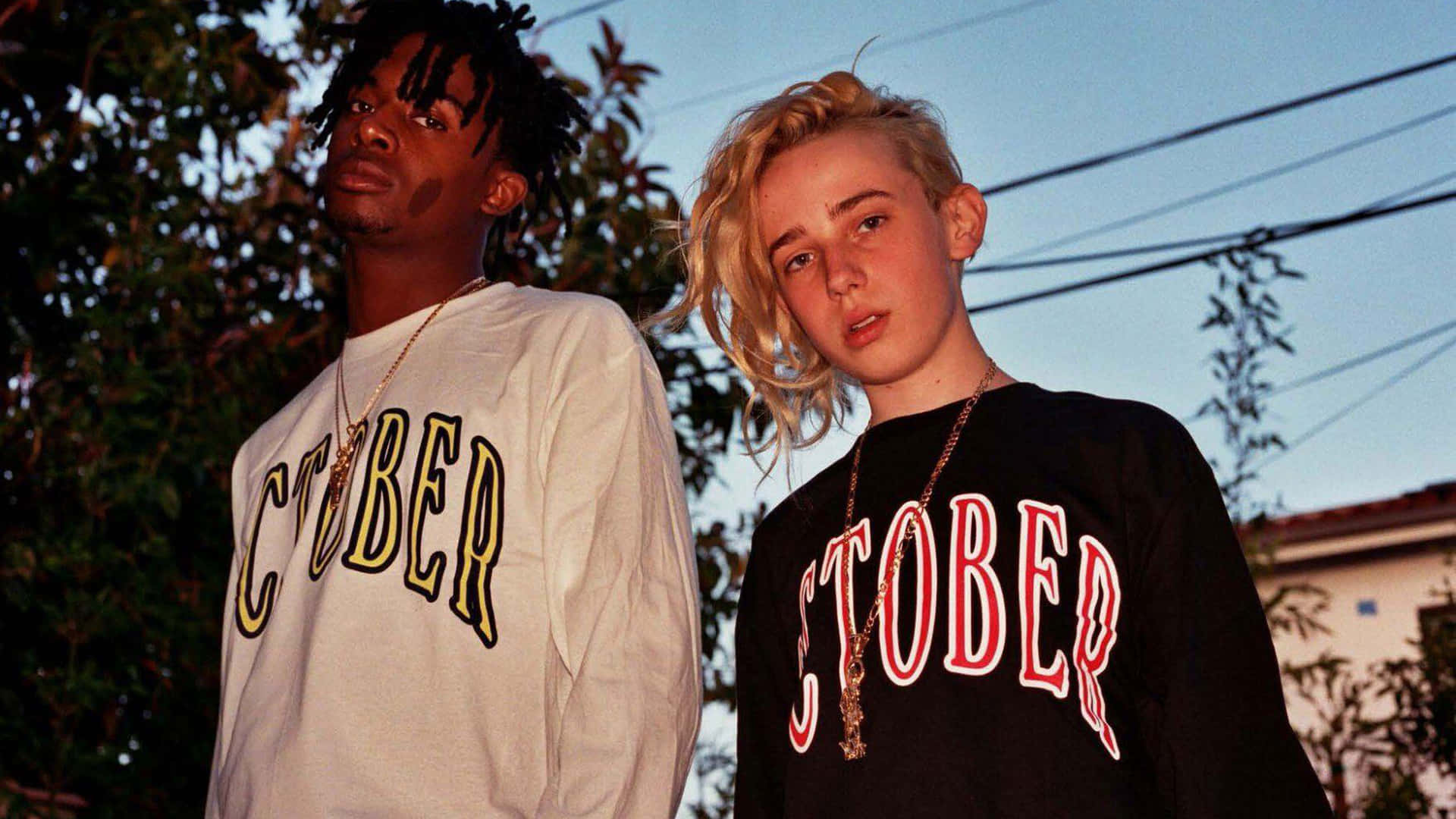 Playboi Carti HD 桌布 壁紙 — 兩名身穿'OCTOBER'連帽衫的年輕人站在暮色天空下，背後有樹木和電線。