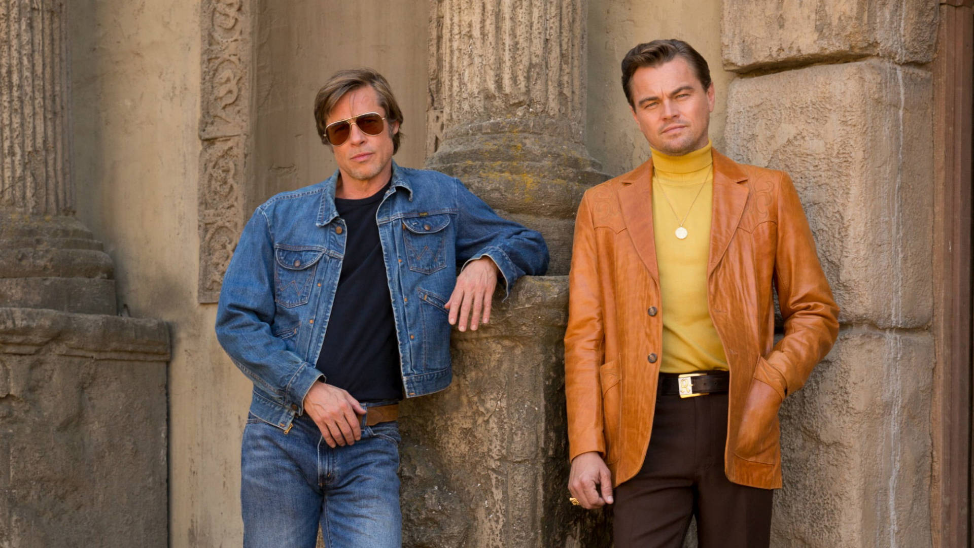 Hollywood Movie 桌布 壁紙 — 兩名身穿70年代風格服裝的男子在明亮的日光下倚靠著古老的石柱。