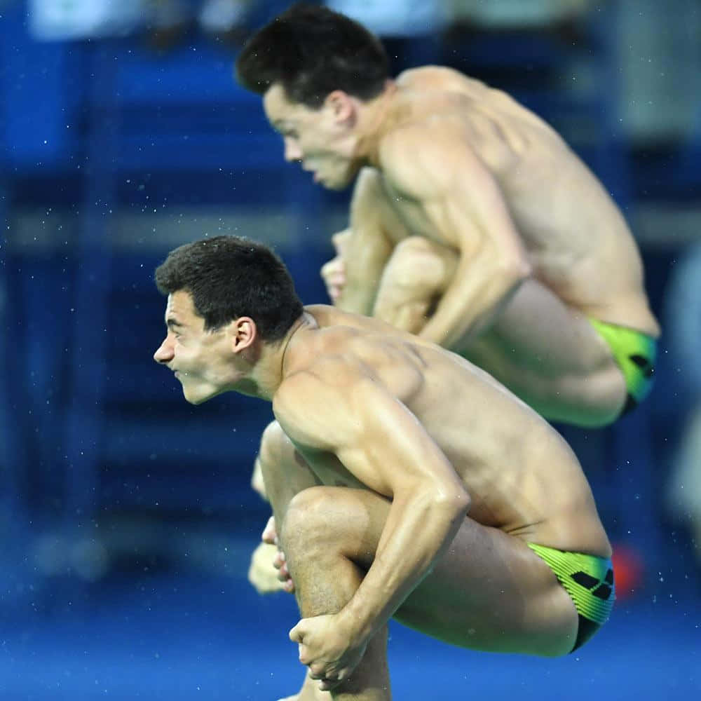 diving 桌布 壁紙 — 兩名身穿亮綠色泳褲的男性潛水員在藍色水滴旋轉的泳池上方半空中蹲伏。