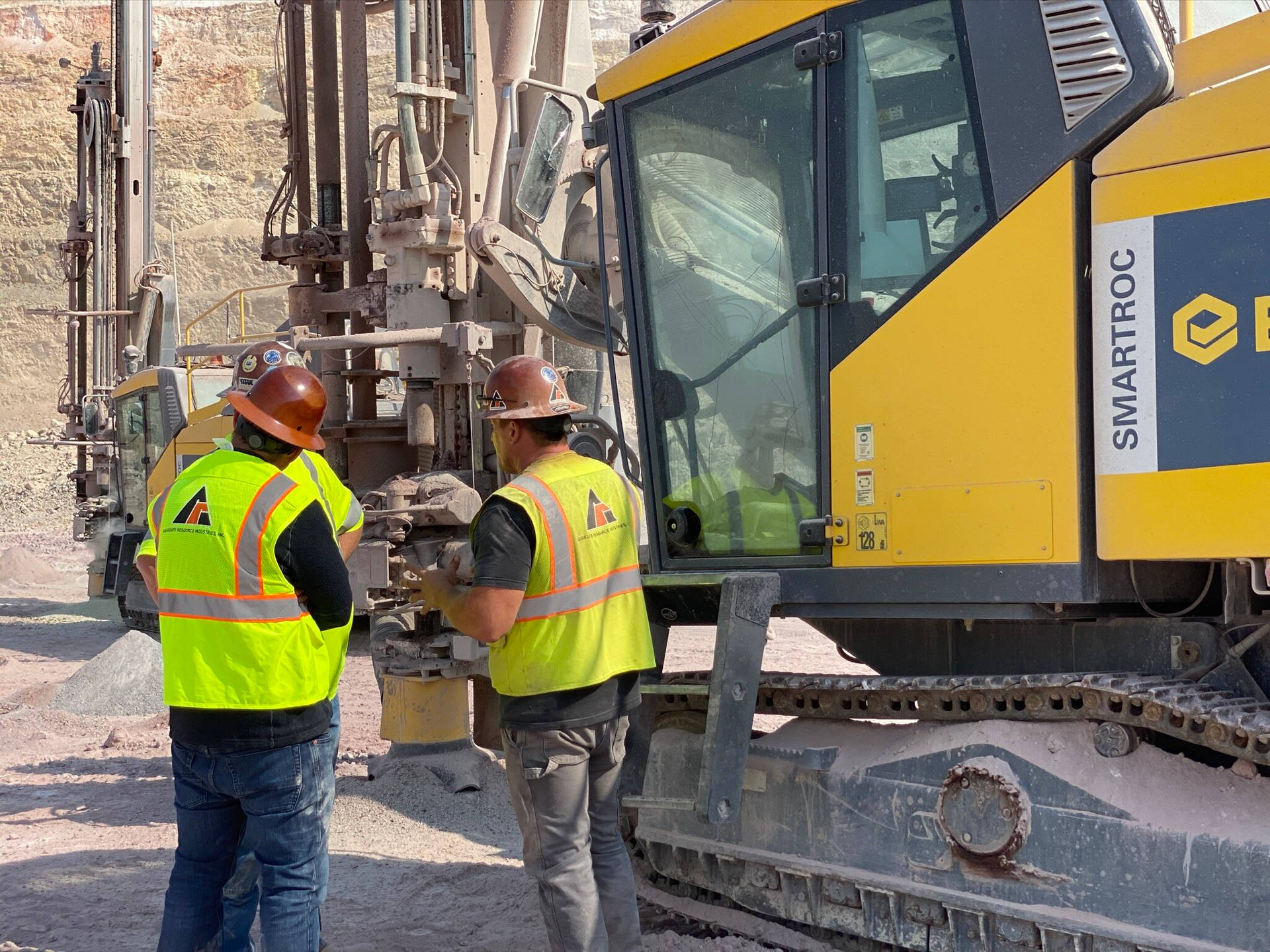 construction 桌布 壁紙 — 兩名身穿亮黃色背心的建築工人在陽光明媚的岩石採石場中，檢查一台大型黃色SmartROC鑽機旁的機械。