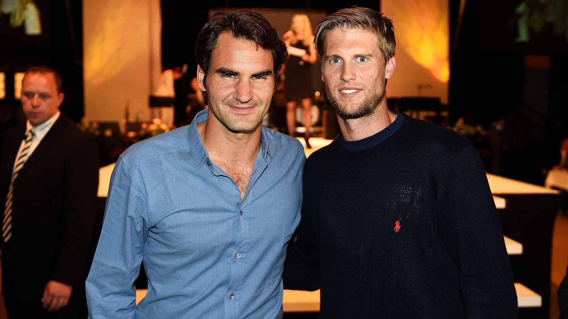 Tennis 桌布 壁紙 — 兩名身穿休閒服裝的男子在室內活動中拍照，環境光線溫暖，背景人物模糊。