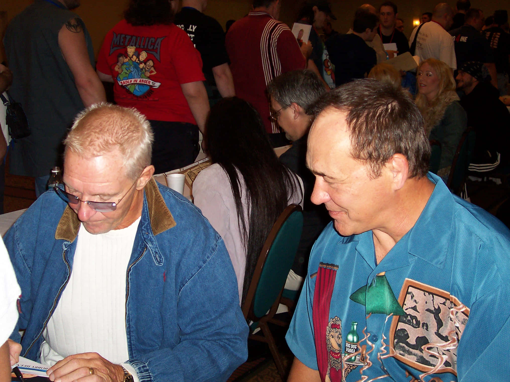Bobby Heenan 桌布 壁紙 — 兩名身穿休閒服裝的男子在擁擠的室內活動中並排而坐，其中一人正在簽名，另一人微笑著觀看。