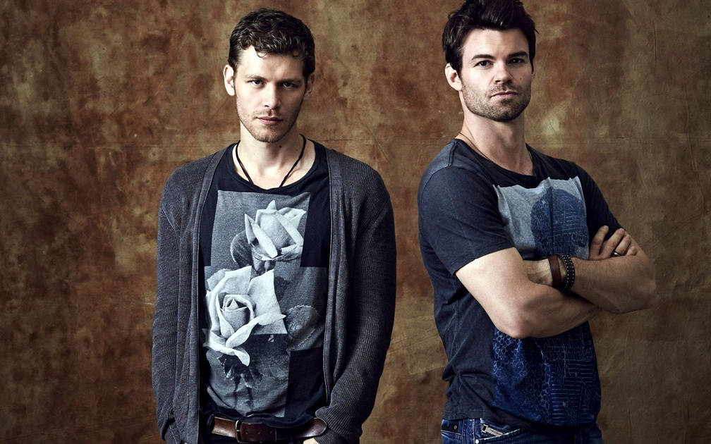 elijah mikaelson 桌布 壁紙 — 兩名身穿休閒服裝的男子自信地站在有紋理的棕色背景前，表情嚴肅。