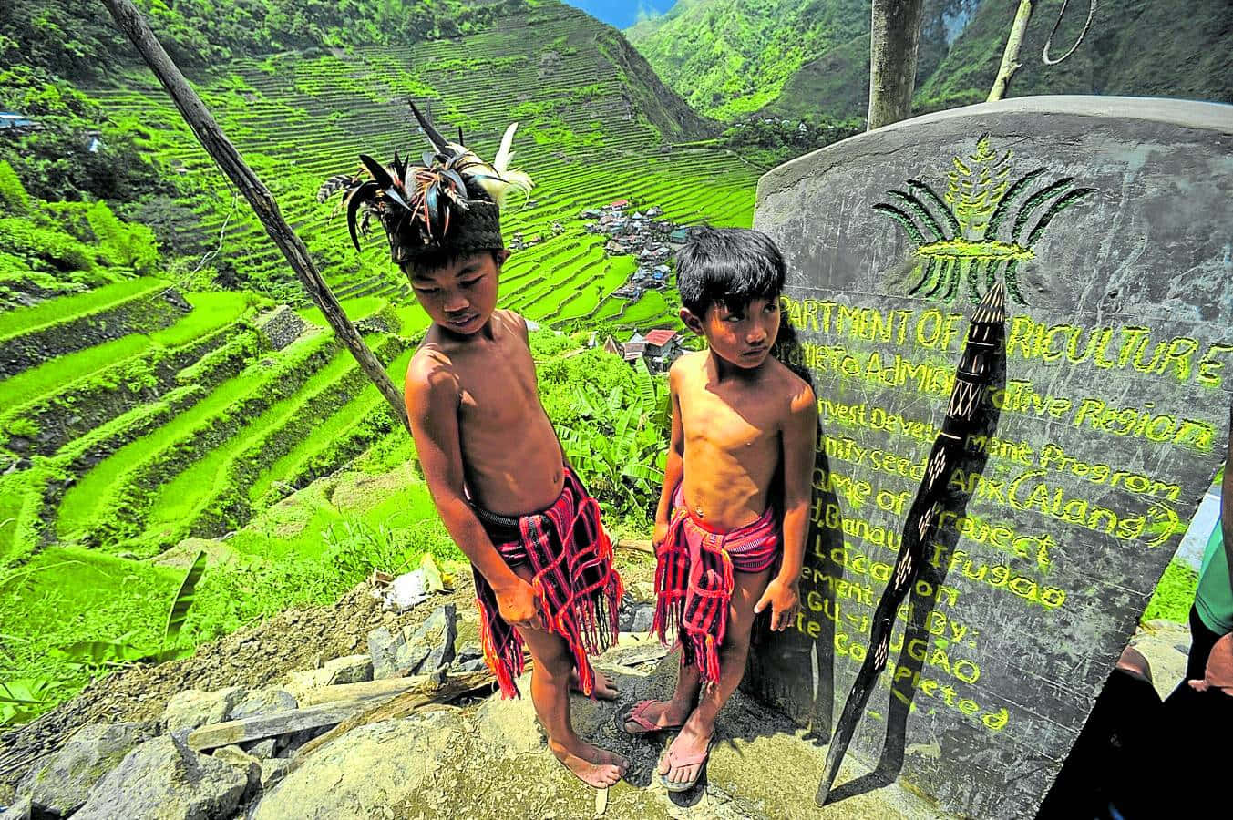 山景 桌布 壁紙 — 兩名身穿傳統服飾的年輕男孩站在政府標誌旁，俯瞰山區中充滿活力的綠色梯田。
