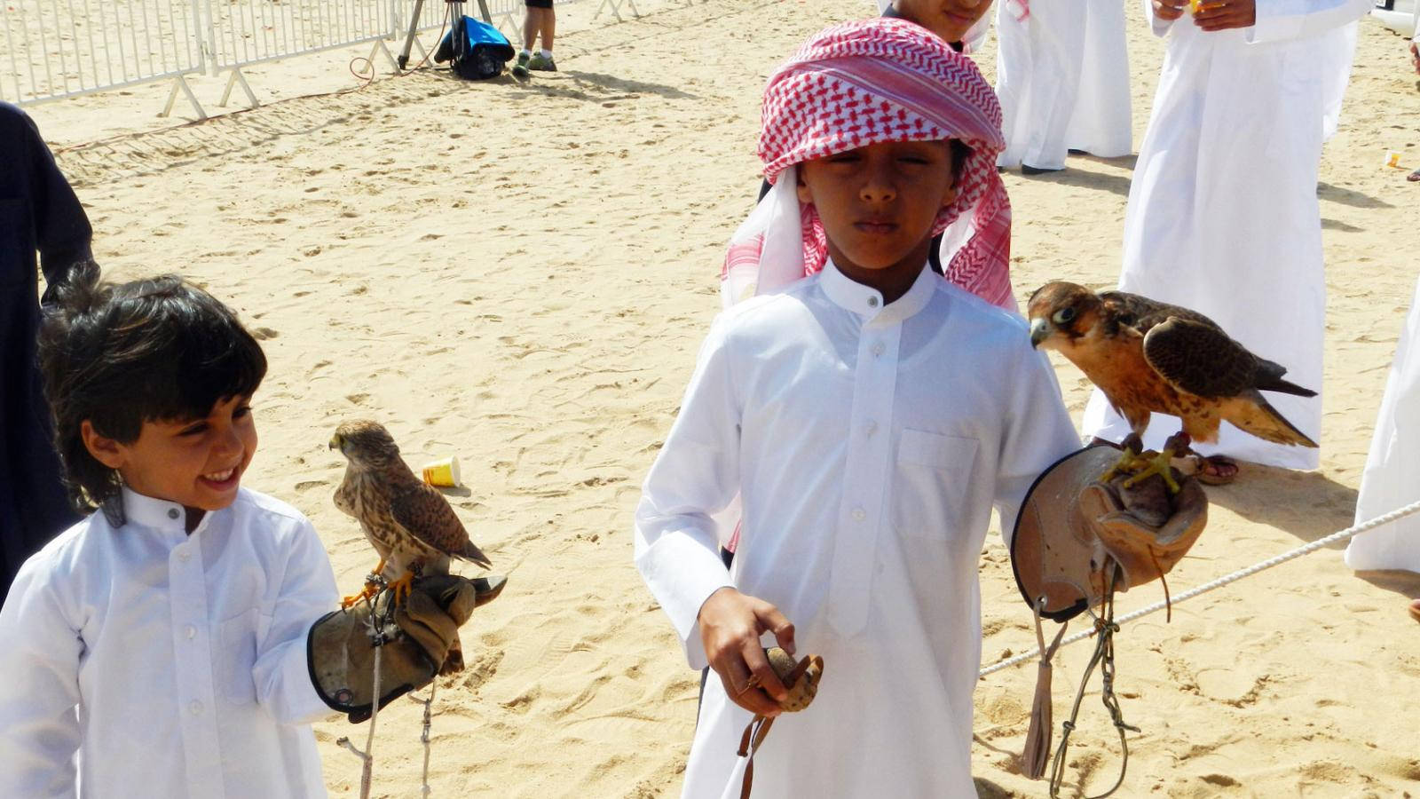 Qatar 桌布 壁紙 — 兩名身穿傳統白色服裝的年輕男孩站在沙灘上，在明亮的日光下，每人手持一隻棲息在戴手套的手上的獵鷹。