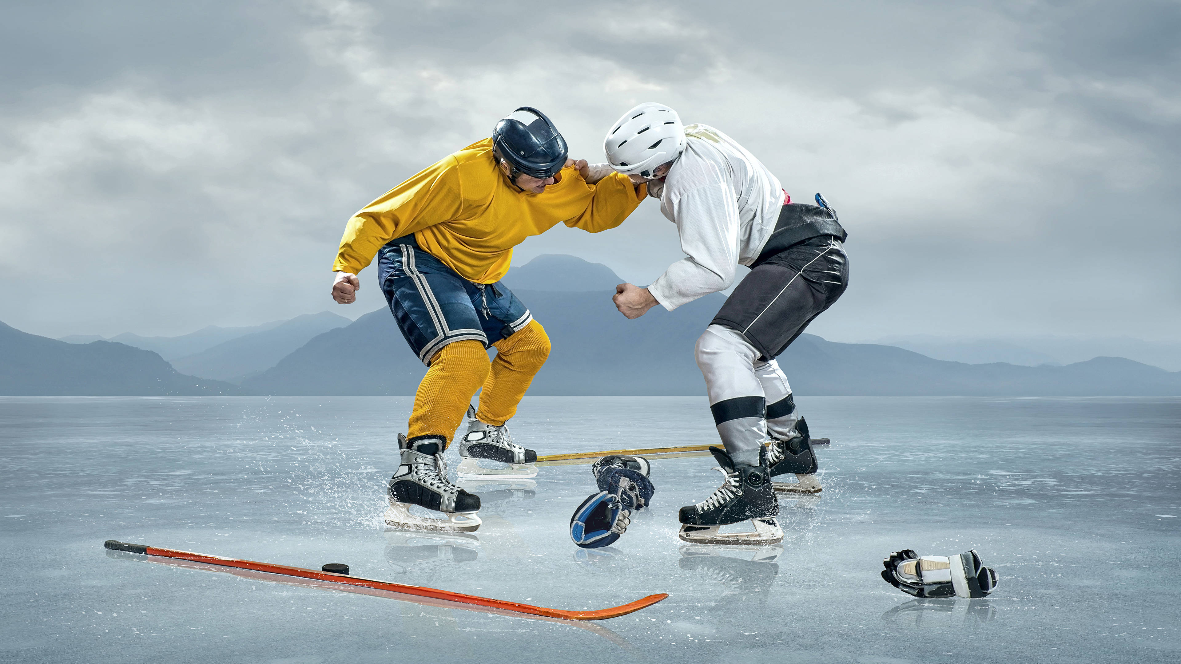 Ice Skating 桌布 壁紙 — 兩名身穿全副裝備的冰球運動員在廣闊的冰凍湖面上激烈纏鬥，背景是霧氣繚繞的山脈，天空多雲。