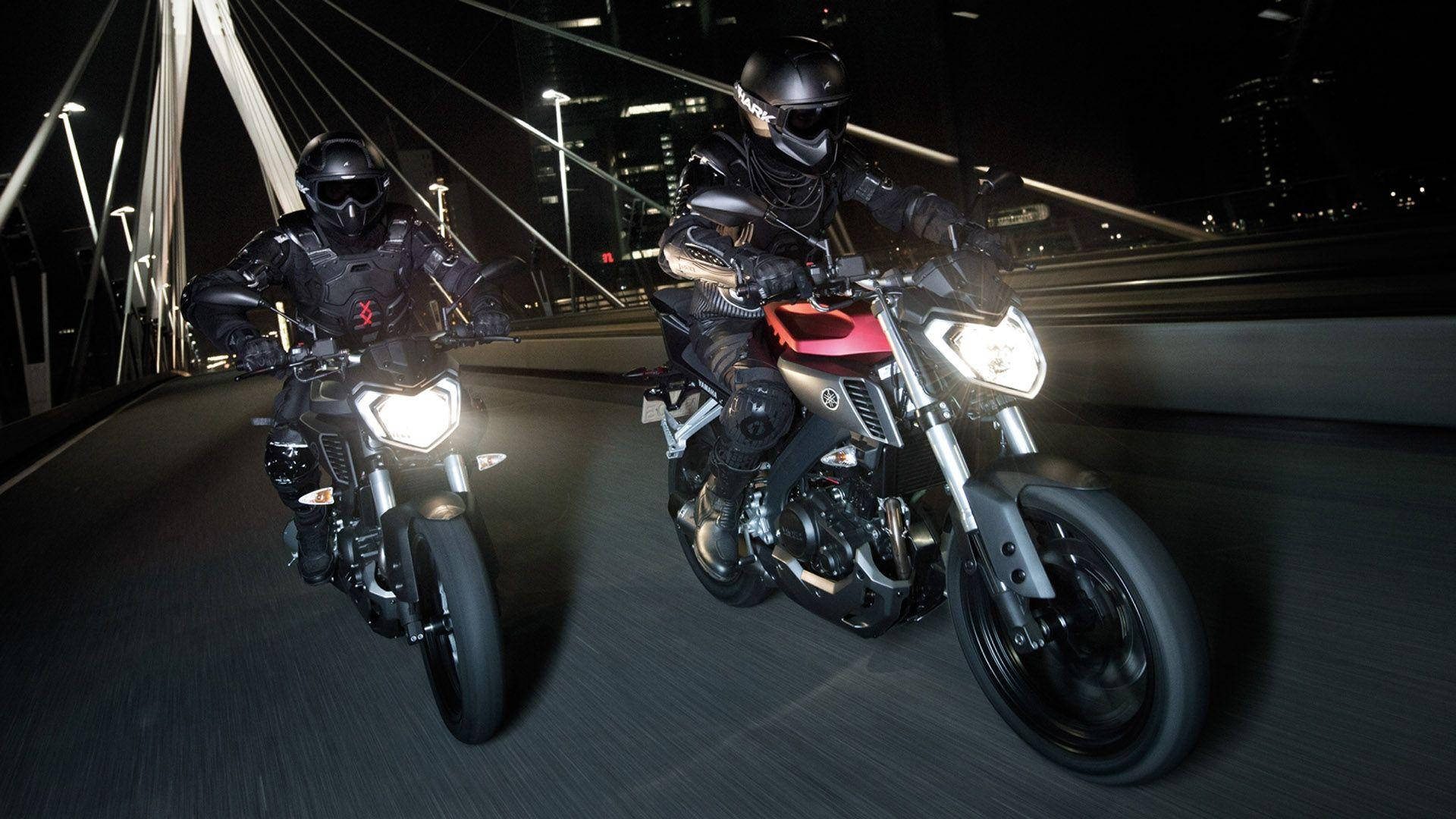 yamaha mt 15 桌布 壁紙 — 兩名身穿全黑裝備的機車騎士在夜間於一座現代化橋樑上競速，身後城市燈光閃爍。