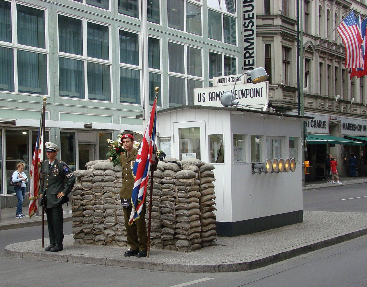 Berlin 桌布 壁紙 — 兩名身穿制服的士兵在柏林歷史性的美軍查理檢查哨站崗，帶著旗幟和沙包屏障。