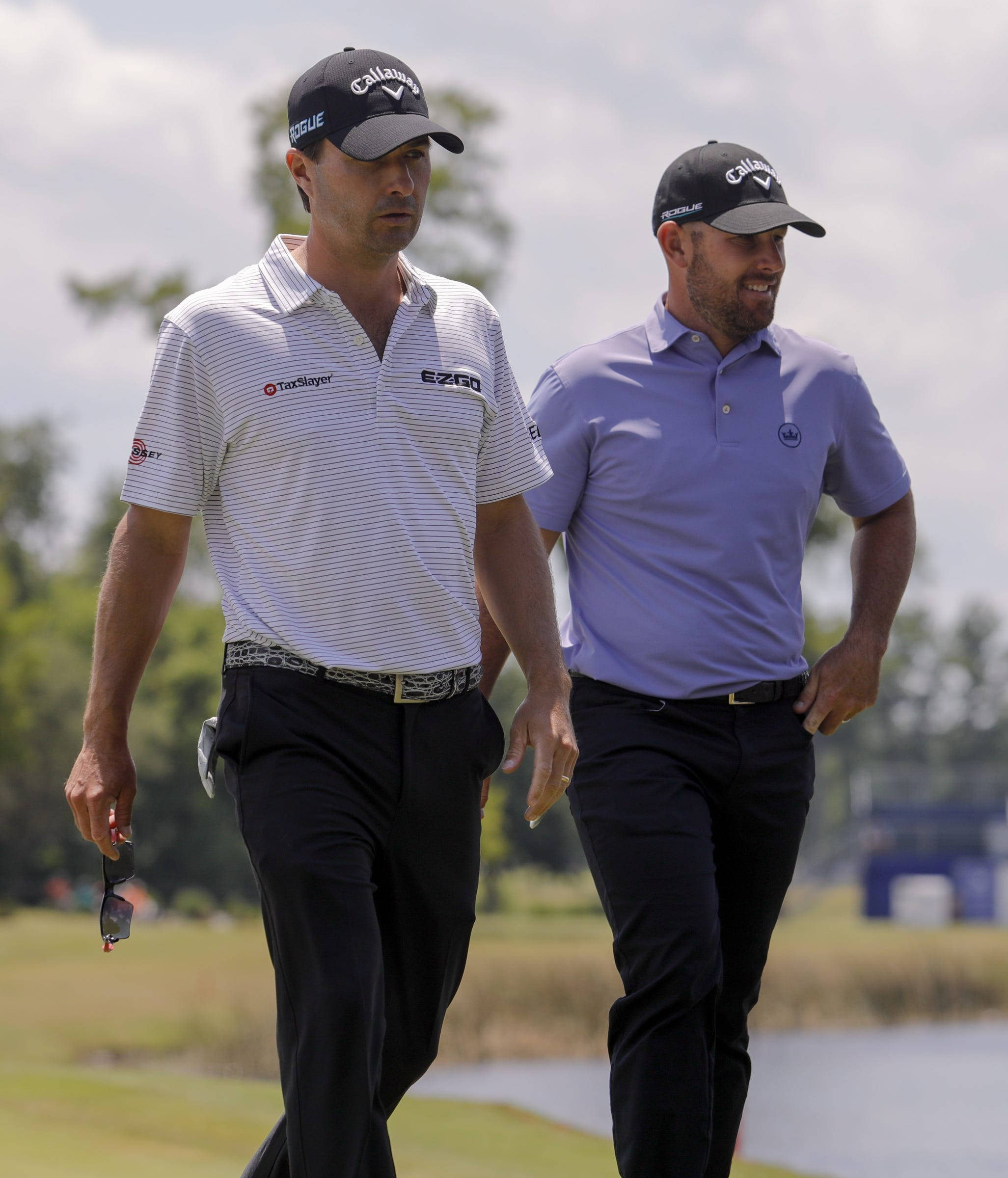 Golfing 桌布 壁紙 — 兩名身穿品牌服裝的男性高爾夫球手在陽光明媚的球場上並肩而行，背景有樹木和水面。