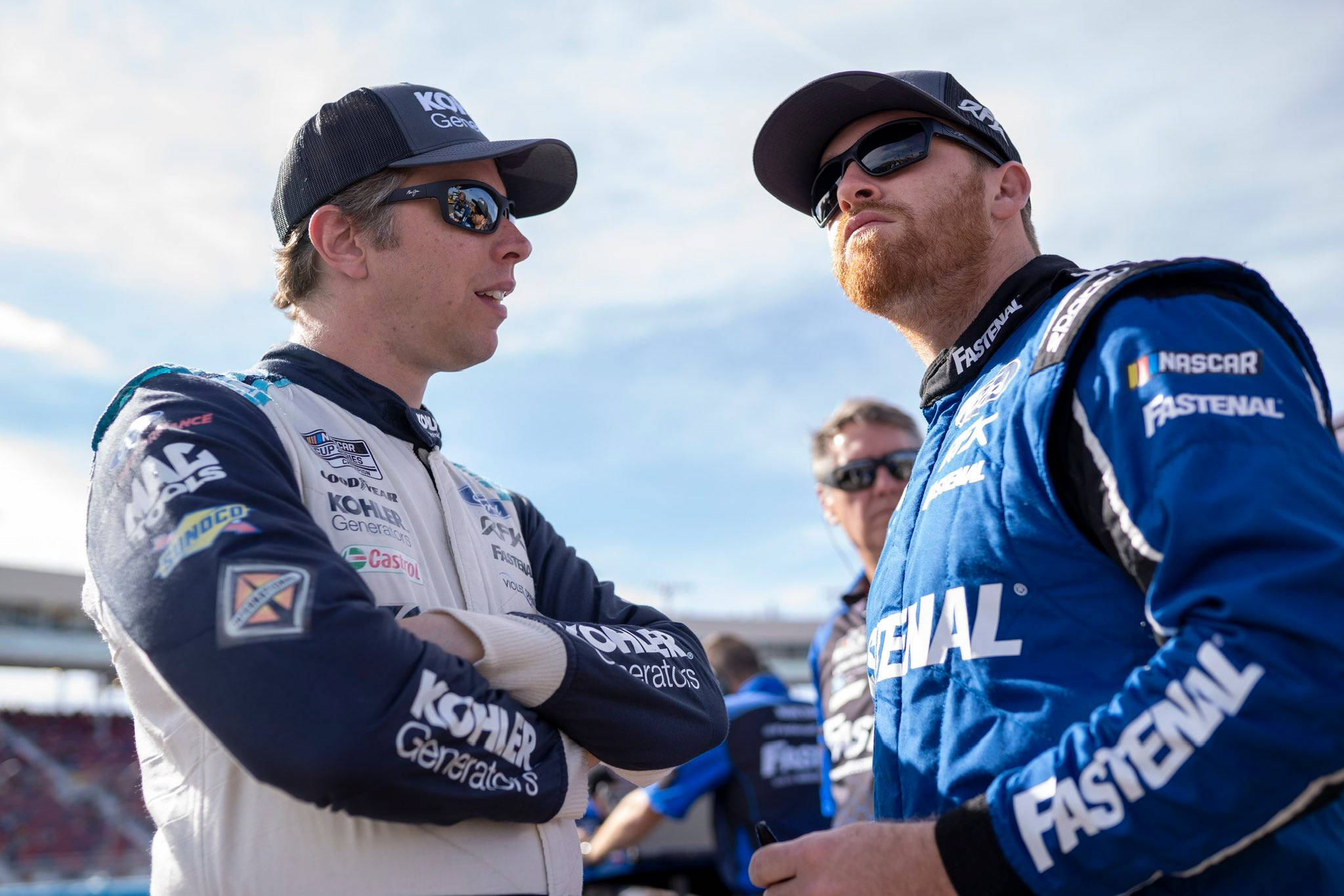 Motorsport 桌布 壁紙 — 兩名身穿品牌賽車服的NASCAR車手在明亮的藍天下於賽道上認真交談。