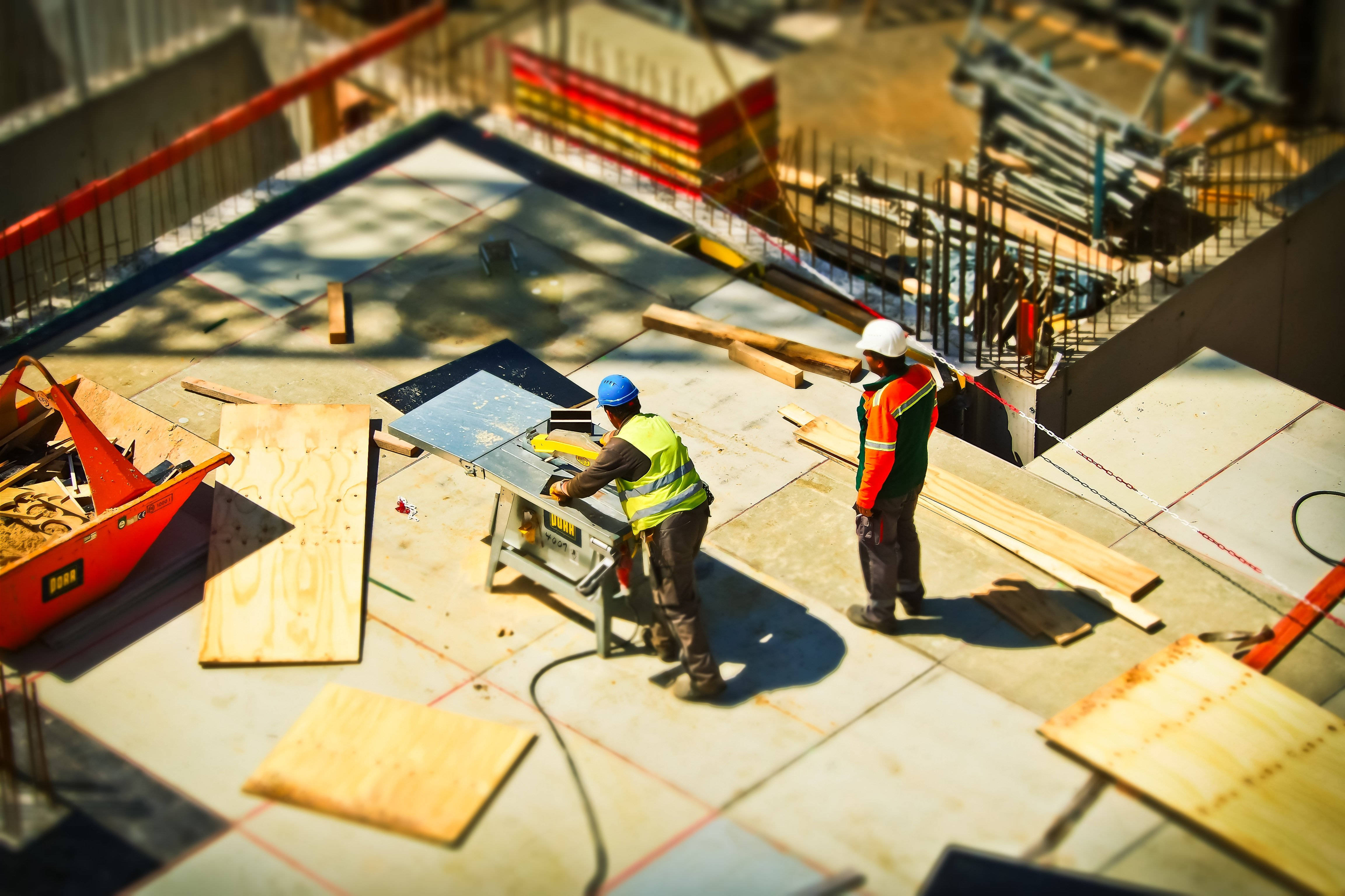 construction 桌布 壁紙 — 兩名身穿安全裝備的建築工人在晴朗天空下，於陽光普照的屋頂上操作檯鋸。
