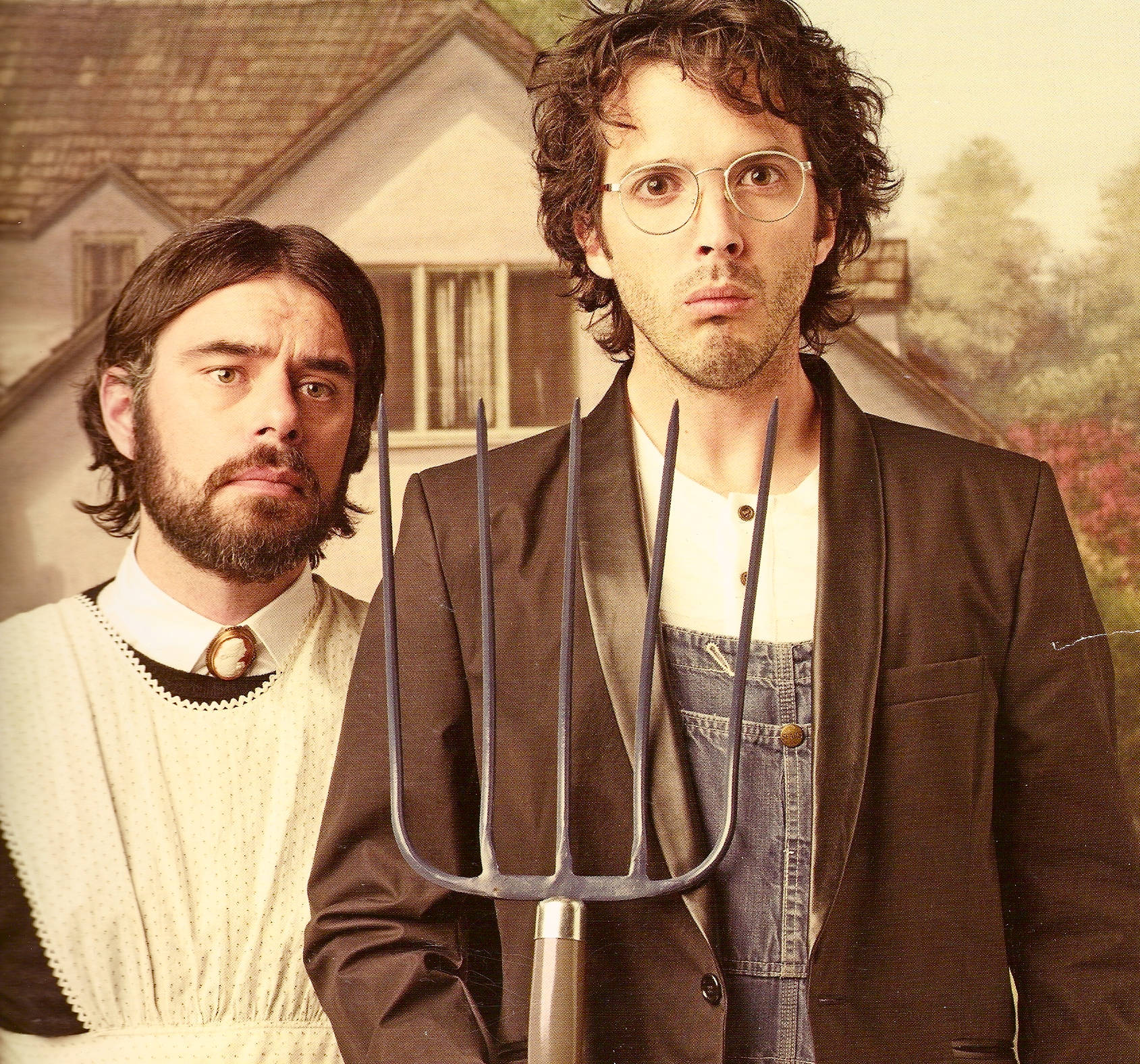 Flight Of The Conchords 桌布 壁紙 — 兩名身穿復古服裝的男子站在一間房子前，其中一人手持乾草叉，在溫暖的金色光線下表情嚴肅。