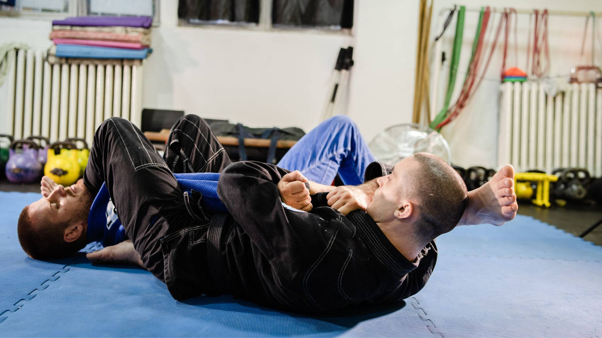 martial arts 桌布 壁紙 — 兩名身穿柔道服的男子在健身房的藍色軟墊上激烈纏鬥，周圍放滿訓練器材。