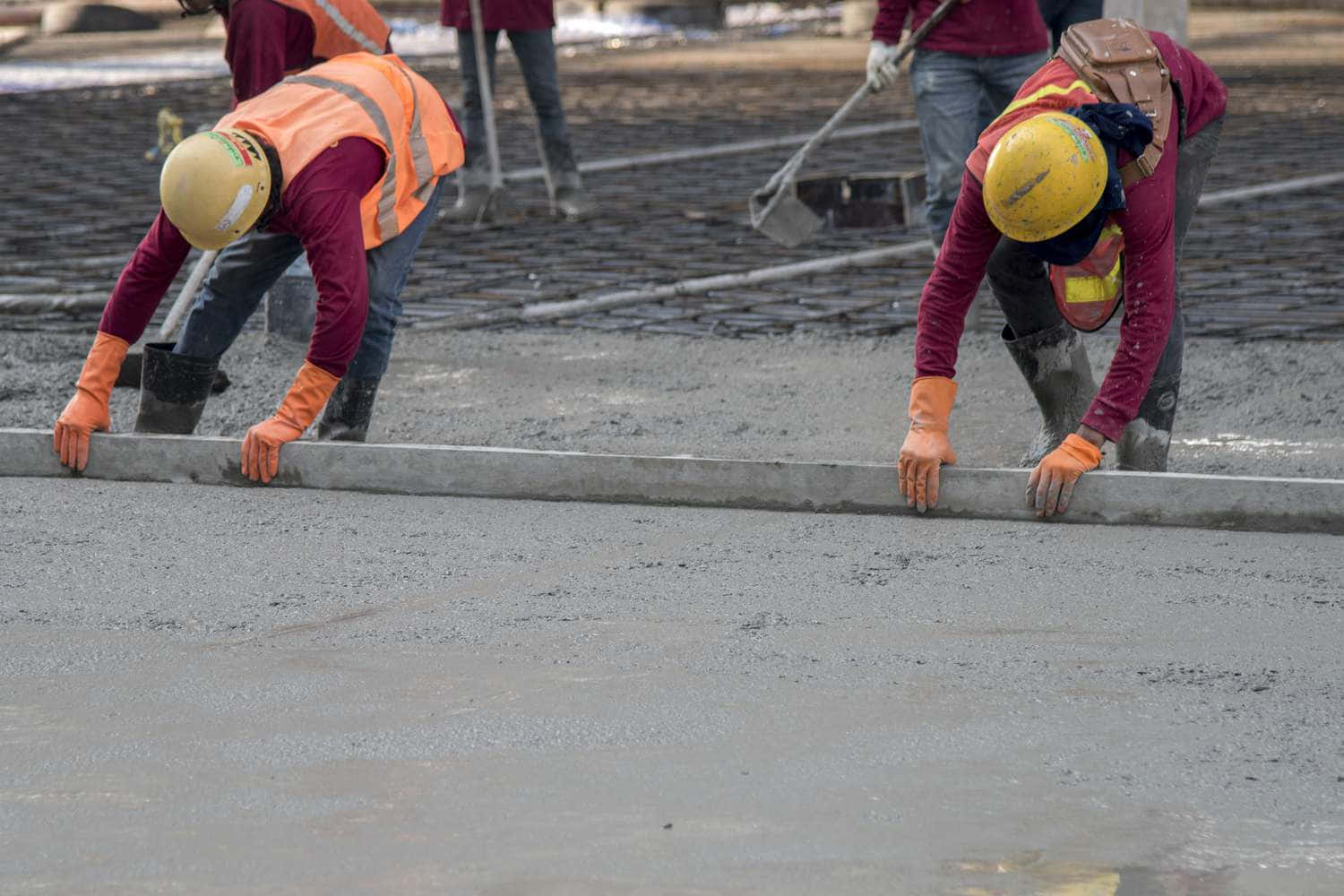 Building 桌布 壁紙 — 兩名身穿橙色背心及黃色頭盔的建築工人在工地用長直尺抹平濕混凝土。