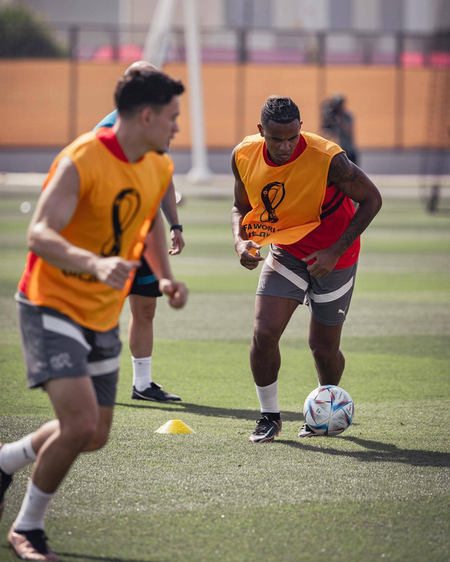 Football Players 桌布 壁紙 — 兩名身穿橙色訓練背心的足球運動員在陽光明媚的綠色草地上專注地盤球繞過角錐。